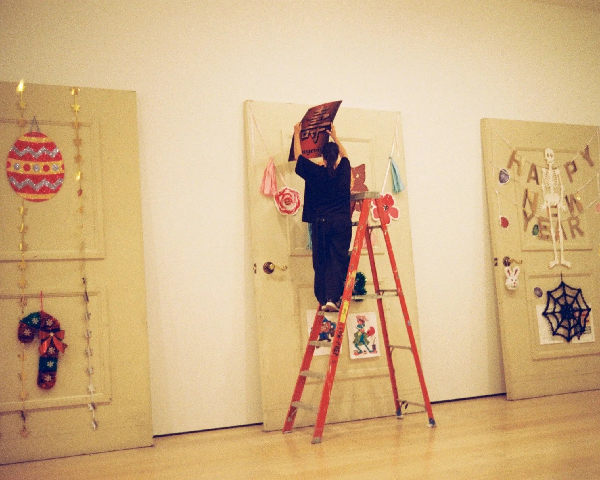 Person standing on a red step ladder hanging a sign on a beige wall decorated with holiday ornaments and artwork.