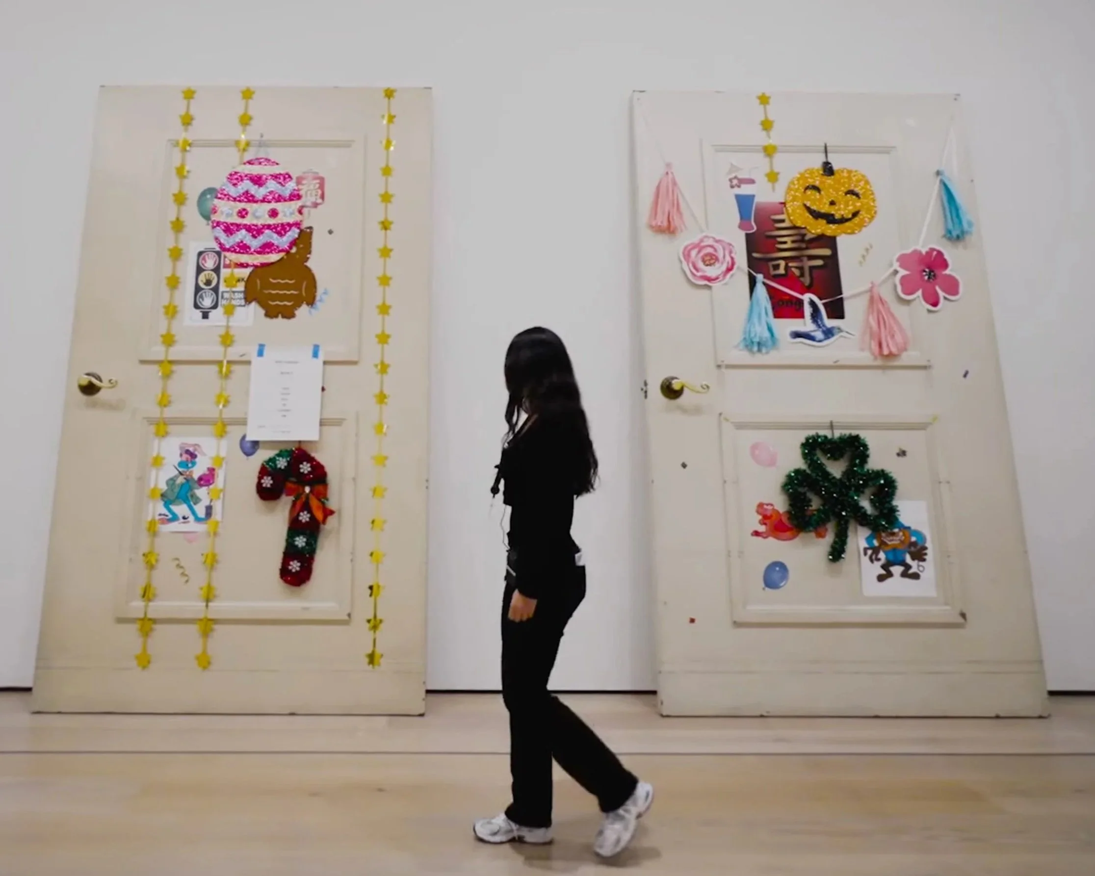 A person dressed in black with white sneakers is performing a dance move indoors, with two decorated doors behind them. The doors are decorated for a festive event with posters, paper flowers, a wreath, and colorful garlands and tassels.