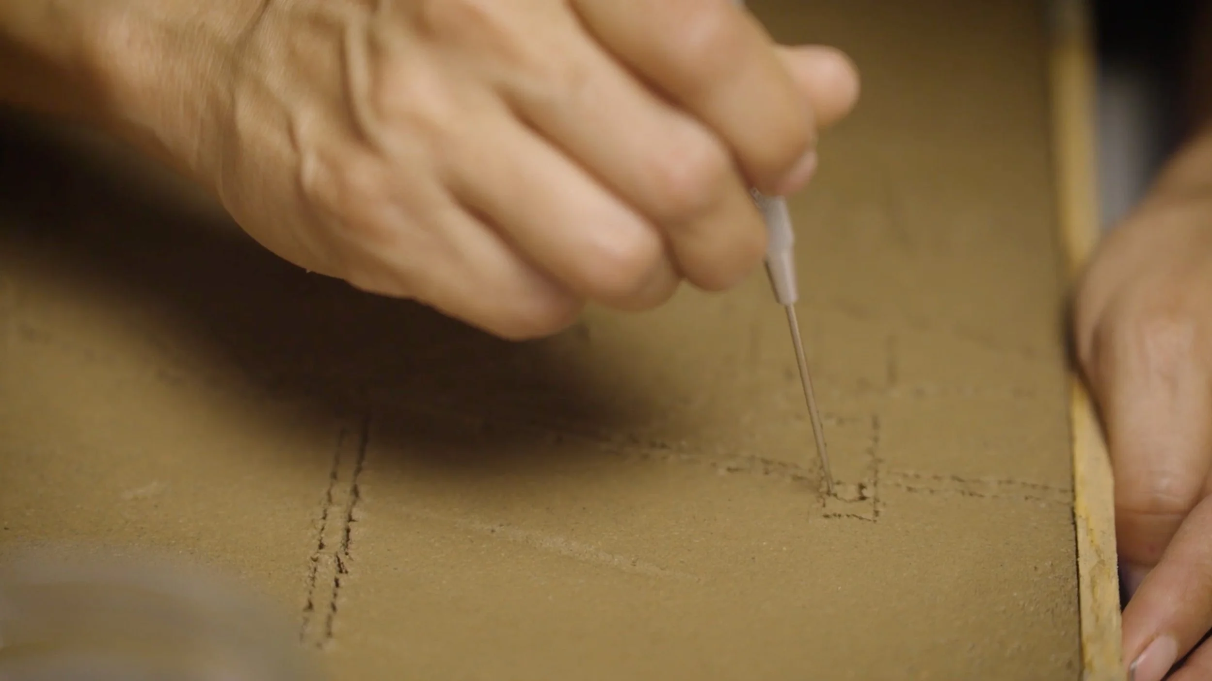 A person's hand is using a needle to sew or stitch along a dashed line on a piece of cardboard or similar material.