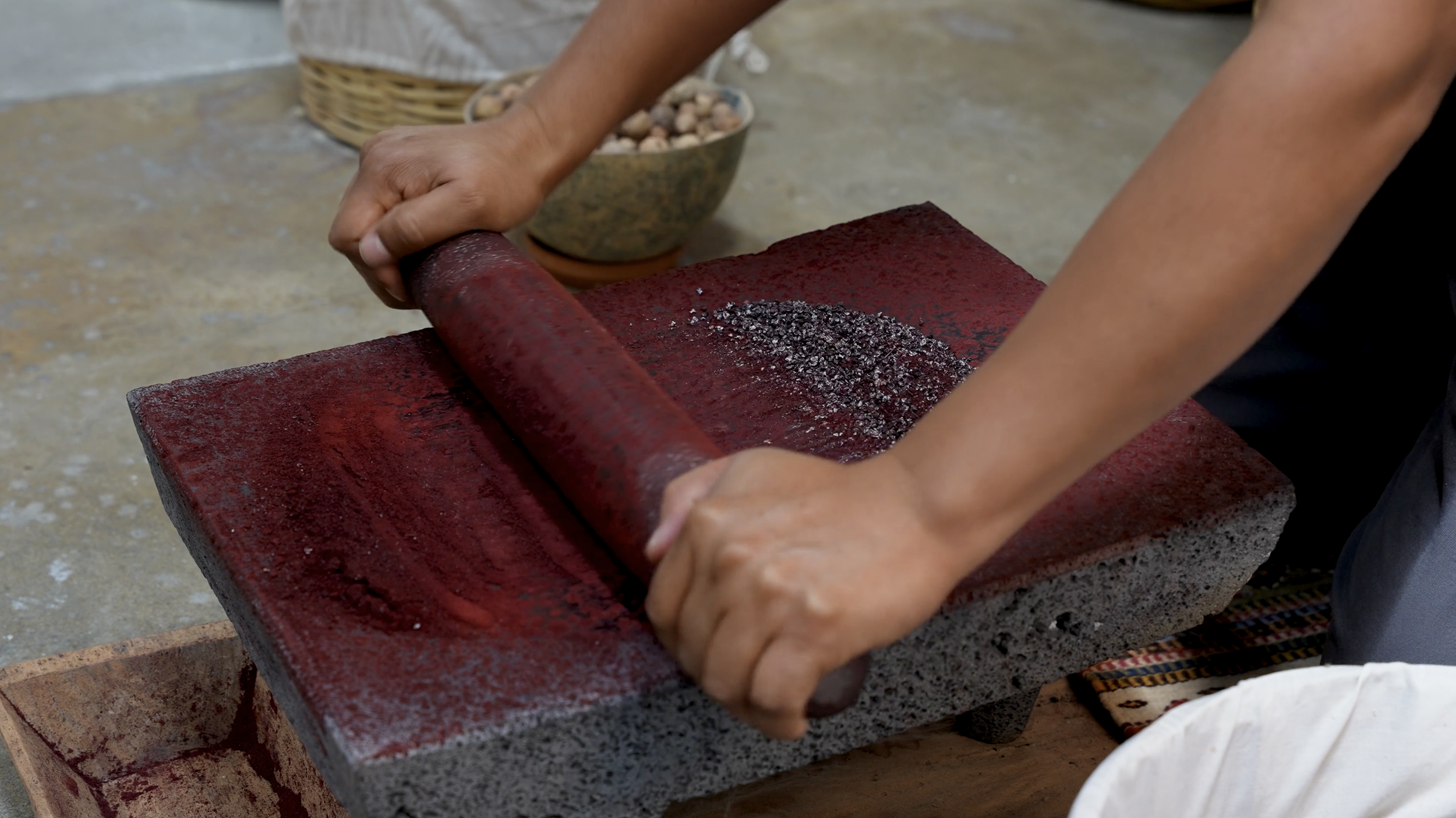 Person using a red mortar and pestle to grind black substance on a red grinding stone, with bowls of small nuts or seeds in the background.