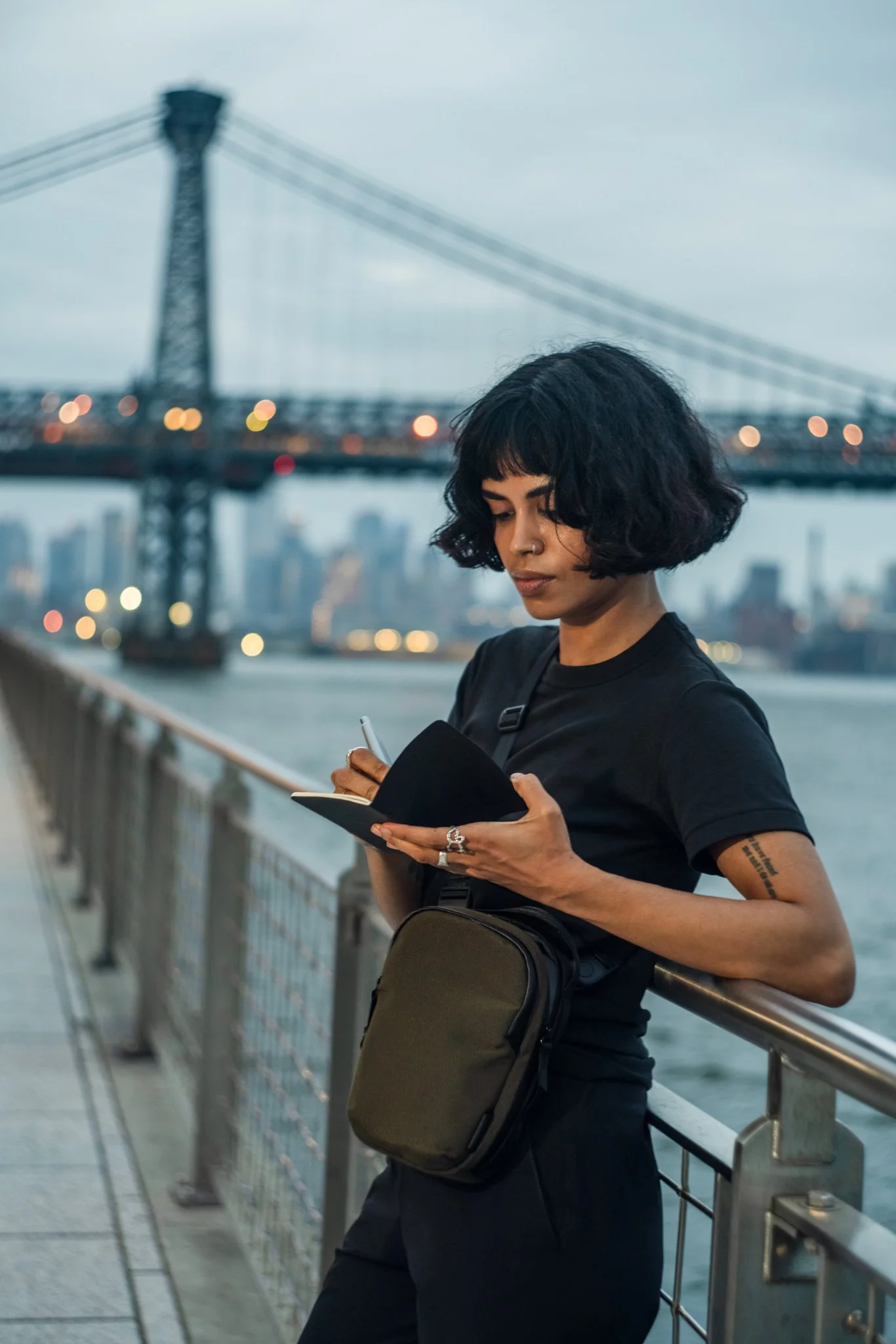 A woman with short curly black hair, dressed in black, is leaning against a railing on a bridge, looking at her phone. She has a nose ring and a tattoo on her left arm. The background shows a city skyline and a large suspension bridge with blurred li