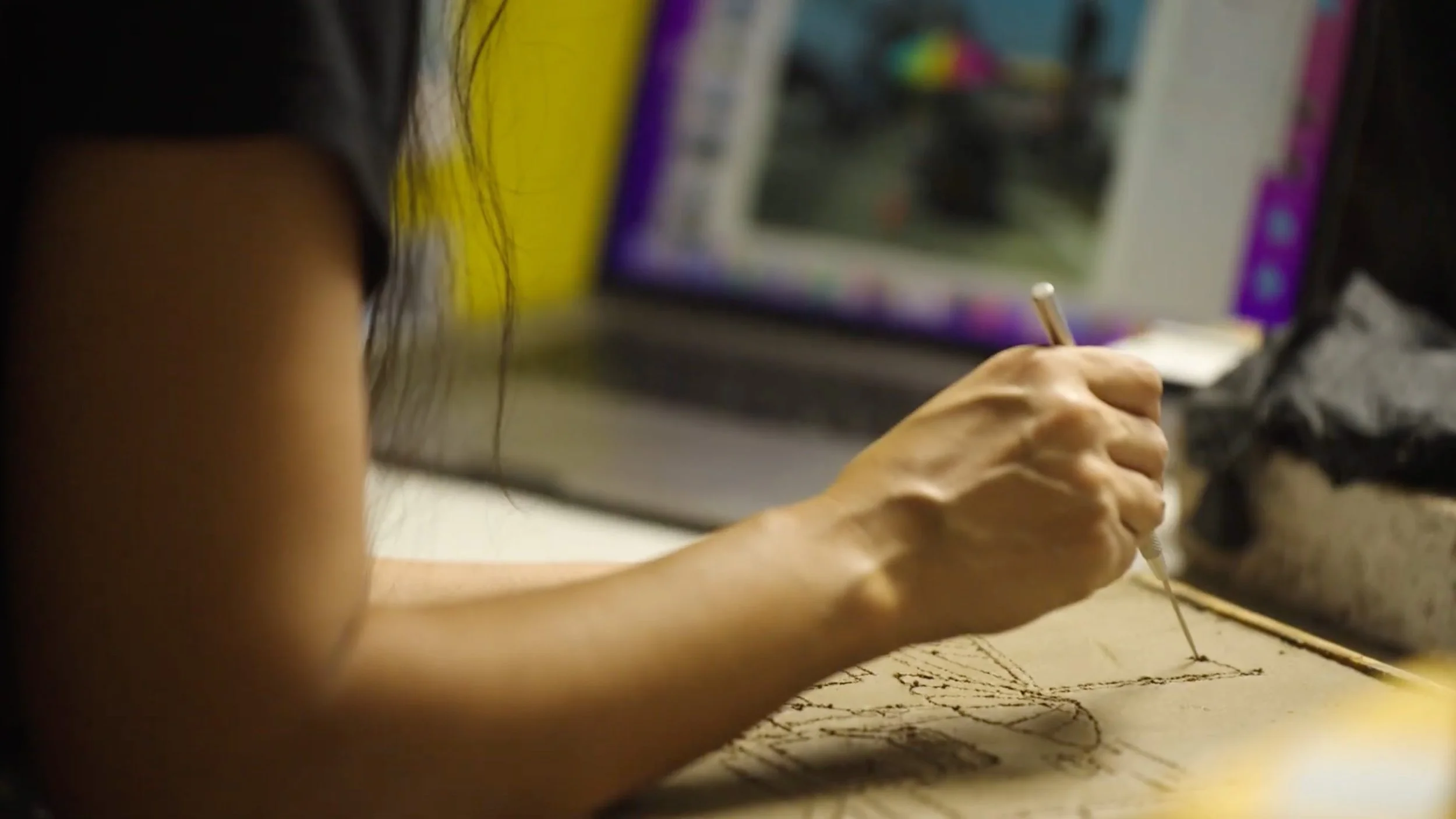 Close-up of a person's hand drawing on a piece of paper with a pencil, with a computer monitor in the background.