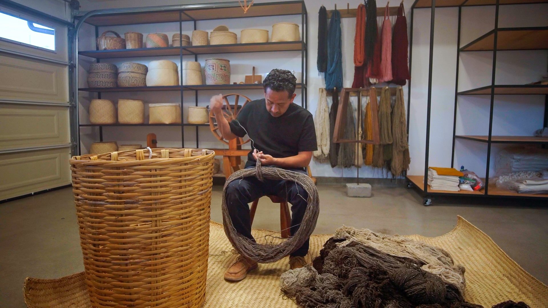 A man sitting on a wooden stool working with natural fibers in a storage room with shelves of woven baskets, colored scarves, and textiles.