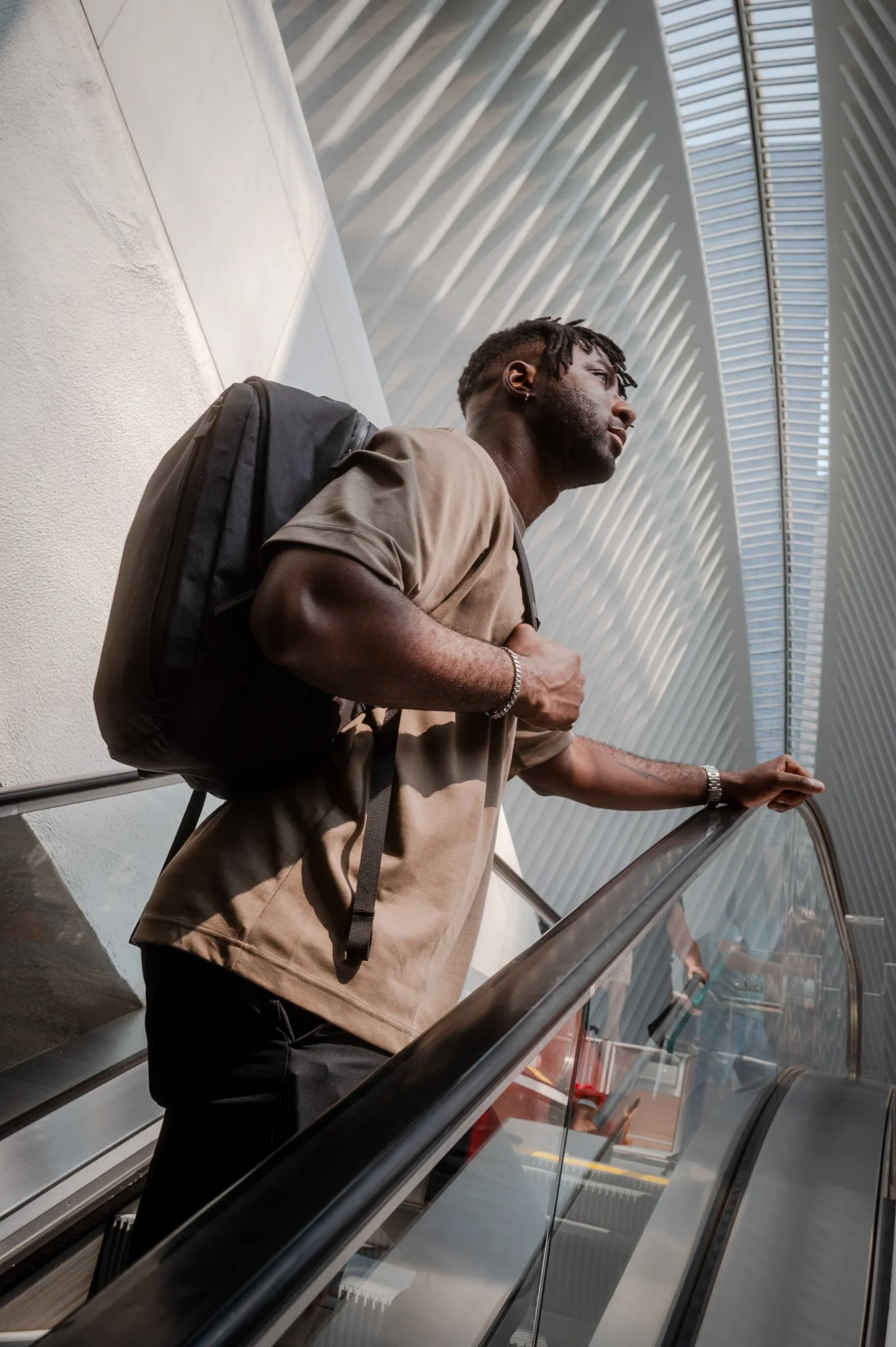 A man wearing a beige t-shirt and carrying a black backpack is standing on an escalator with glass sides, reaching out with his right hand, in an indoor space with modern architectural design featuring a high ceiling with linear patterns and large wi
