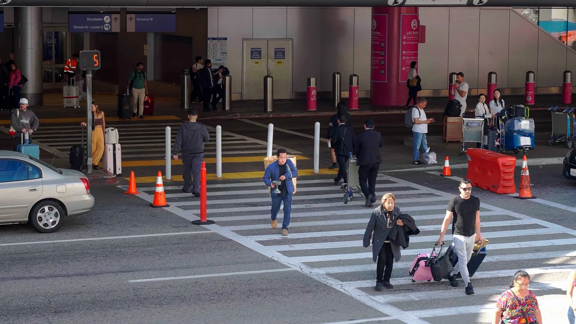 People walking with luggage and carrying bags in front of an airport terminal entrance, with cars parked nearby and various signs indicating check-in and terminal information.