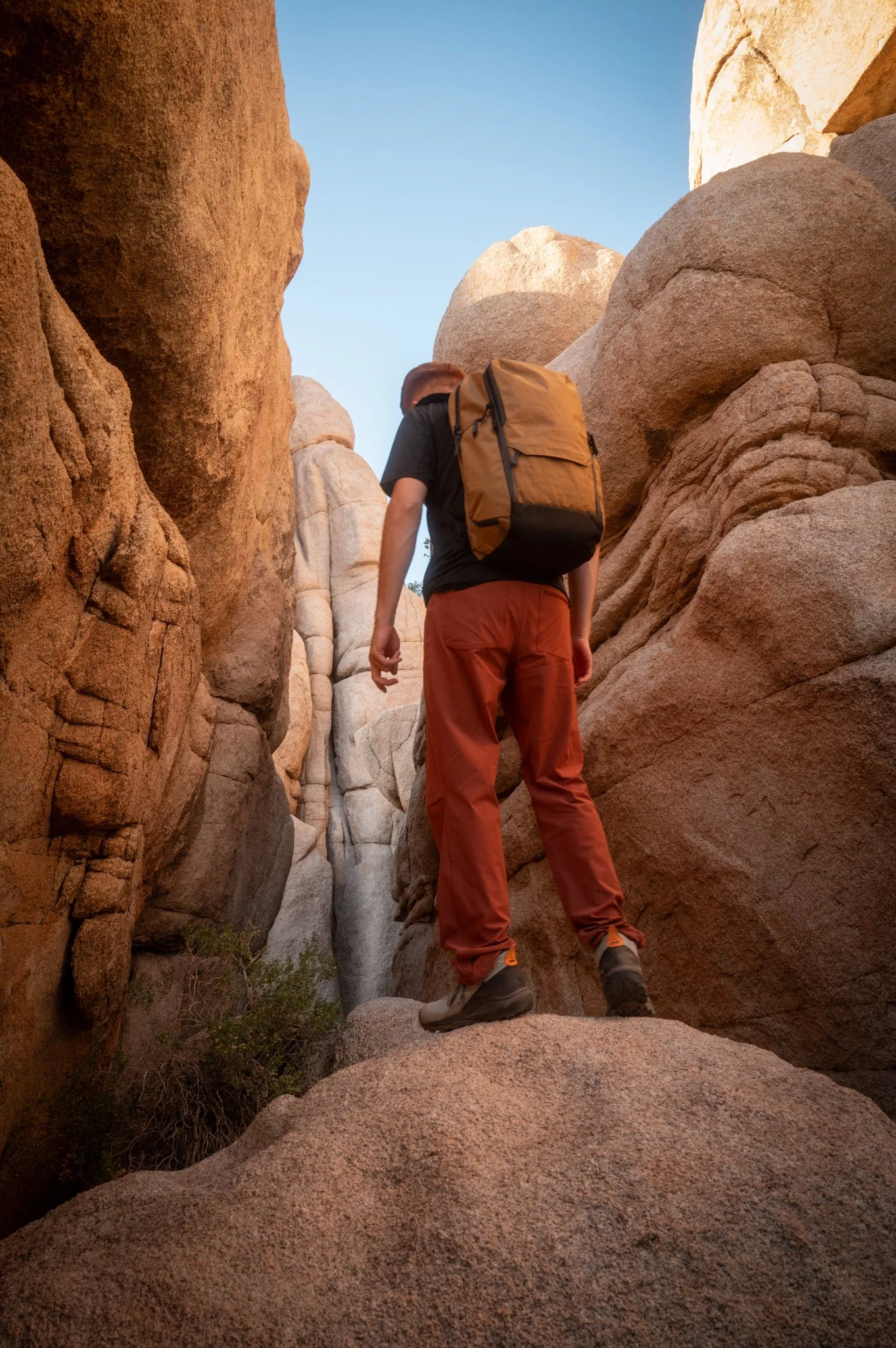 A hiker wearing a black shirt, orange pants, and hiking boots with a tan backpack, standing on large rock formations in a narrow canyon with a clear blue sky overhead.