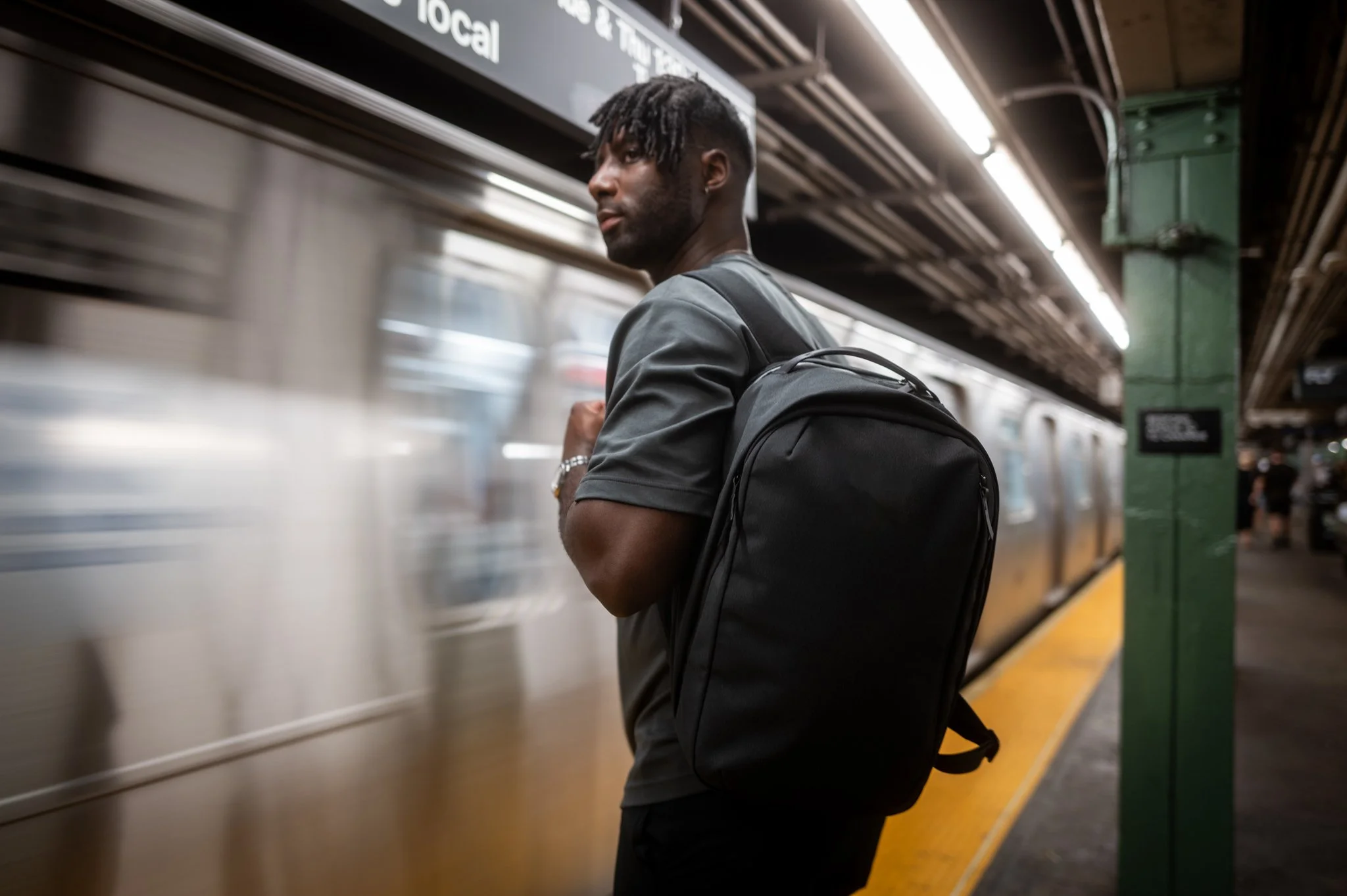 A young man with dreadlocks, wearing a gray shirt and a bracelet, stands on a subway platform with a backpack, looking at a passing subway train.