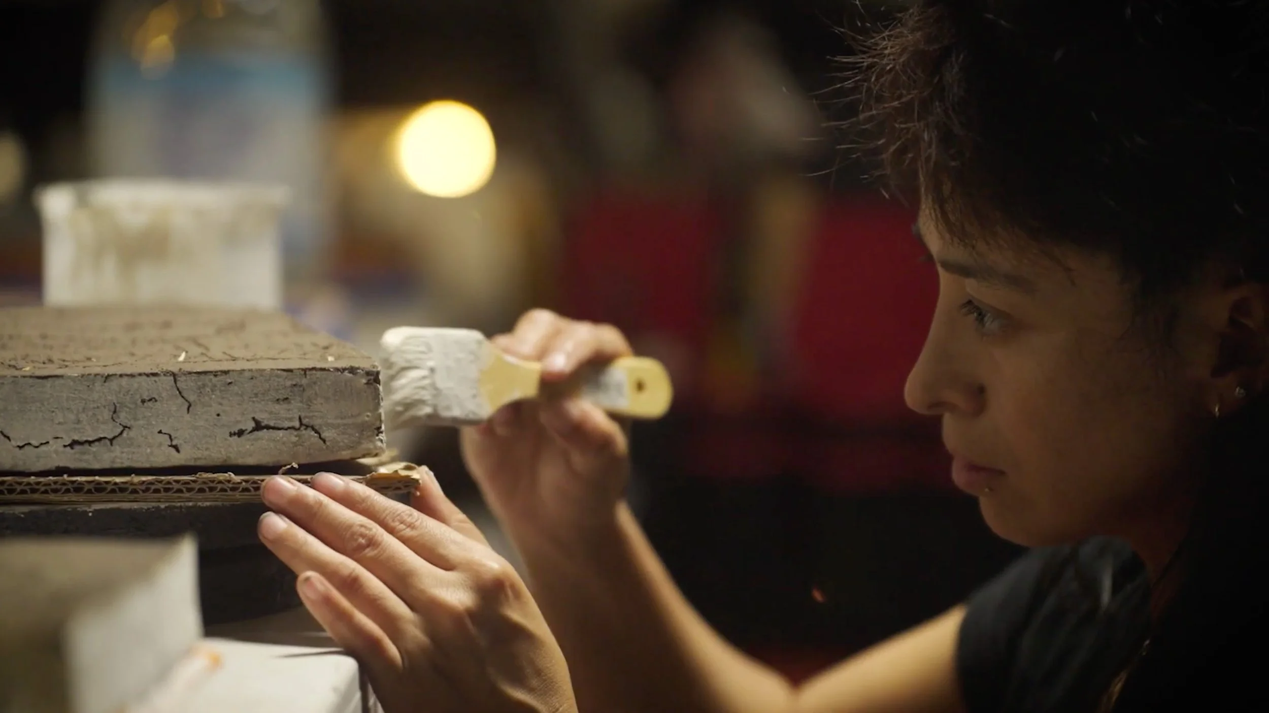 A young person with dark hair carefully painting or detailing a wooden object in a workshop setting.