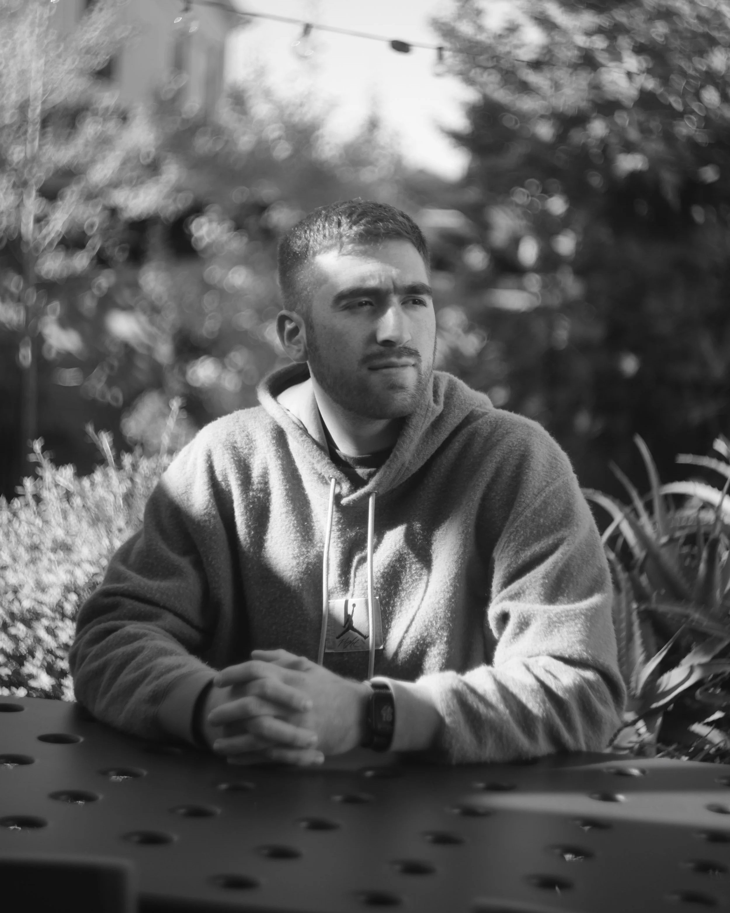 A black and white photo of a young man with short hair and a beard, wearing a hoodie, sitting outdoors in a natural environment with trees and foliage in the background. The photo is taken from behind a perforated surface, possibly a table or bench.