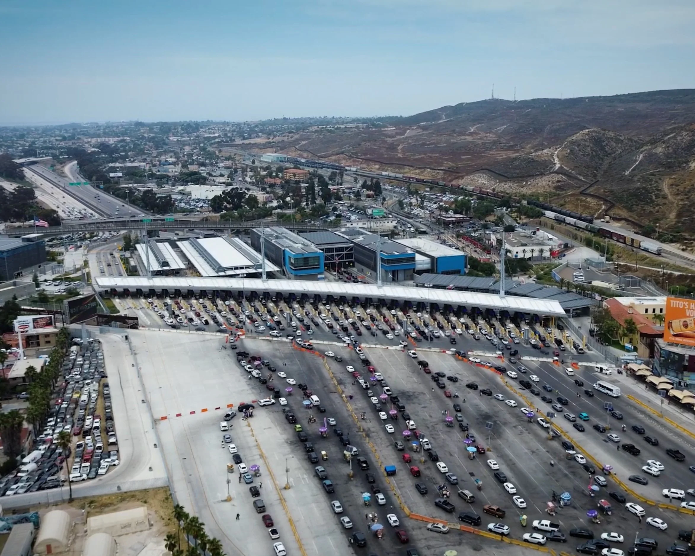 An aerial view of a busy urban area with a large parking lot filled with cars, a highway with vehicles, and a train station with a train on tracks, set against a backdrop of hills and city buildings under a partly cloudy sky.