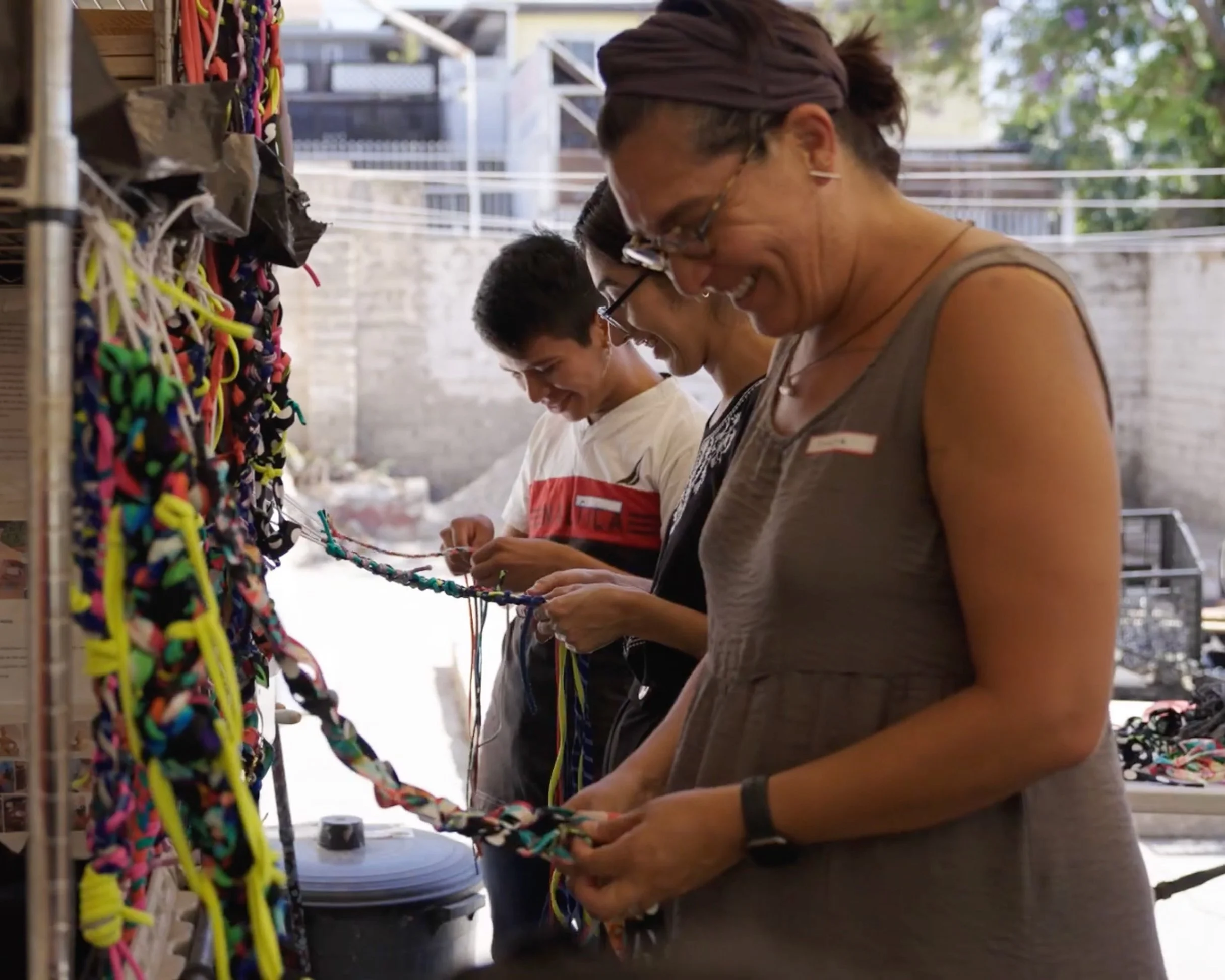 Four people are lookinh down and working on a craft project, with colorful strings hanging on a wall to their left.