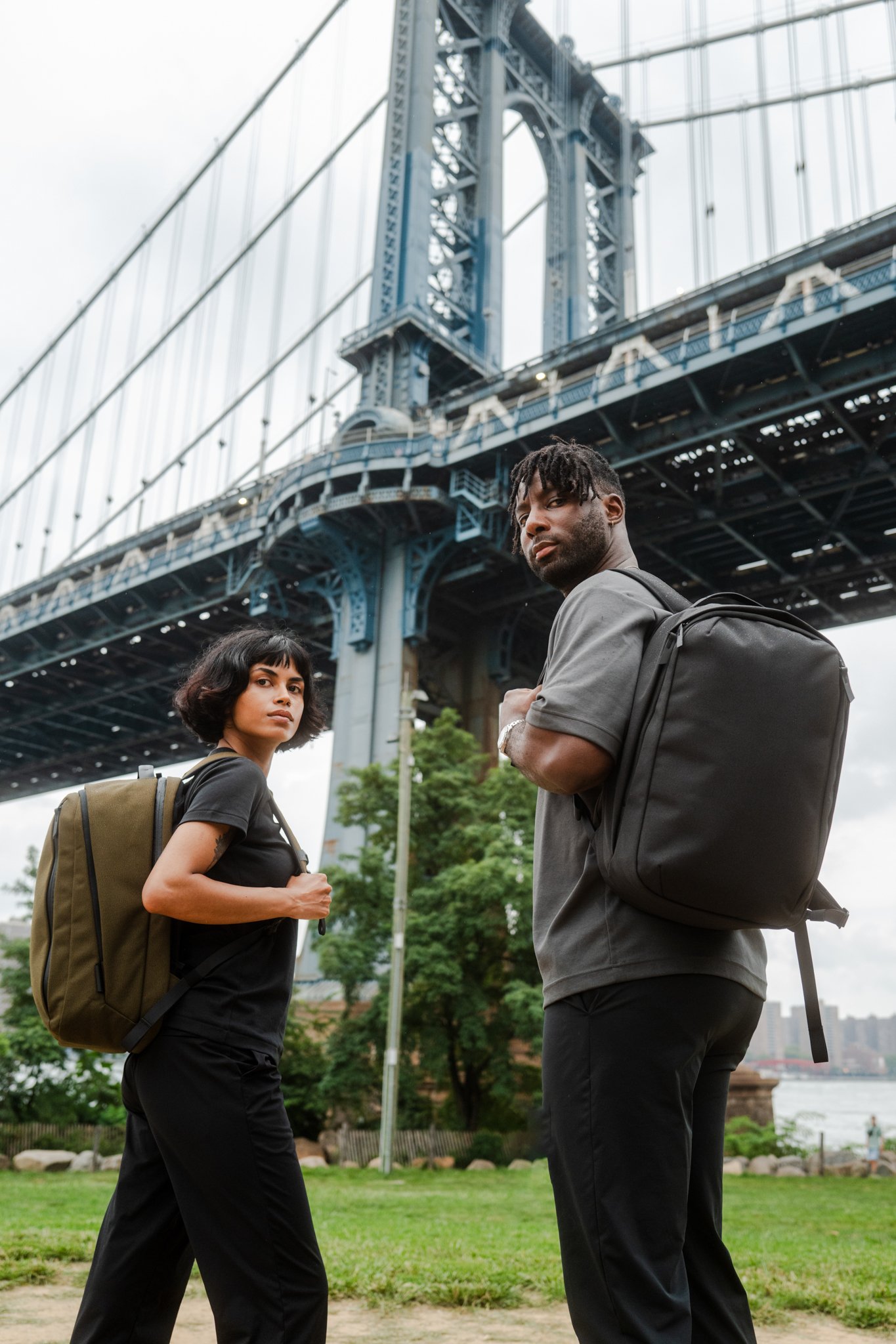 Two young adults with backpacks walking outdoors near a bridge in an urban park.
