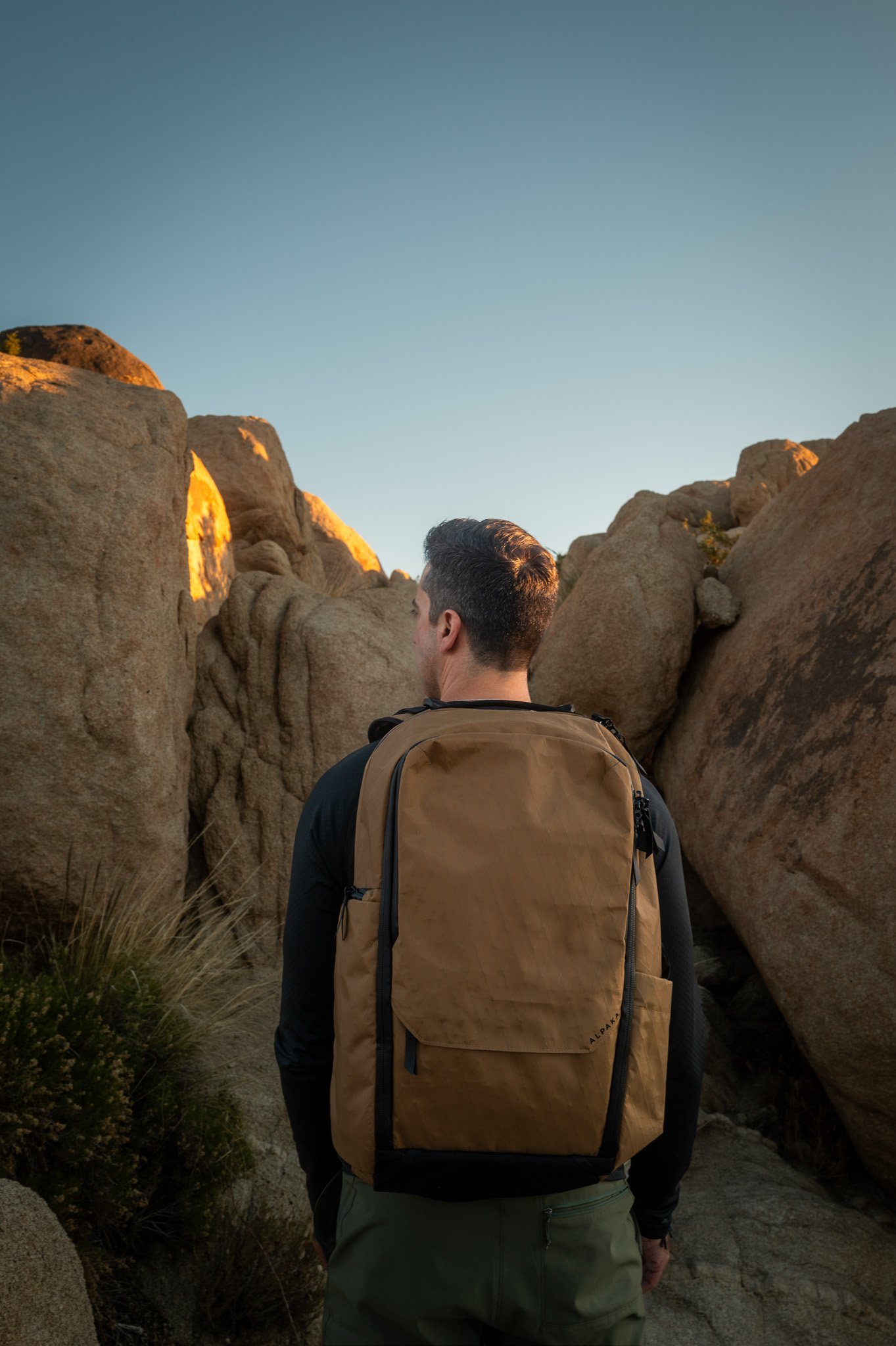 A man with short dark hair, wearing a black jacket, khaki pants, and a tan backpack, walking through a rocky desert landscape with large boulders and sparse vegetation, under a clear blue sky.