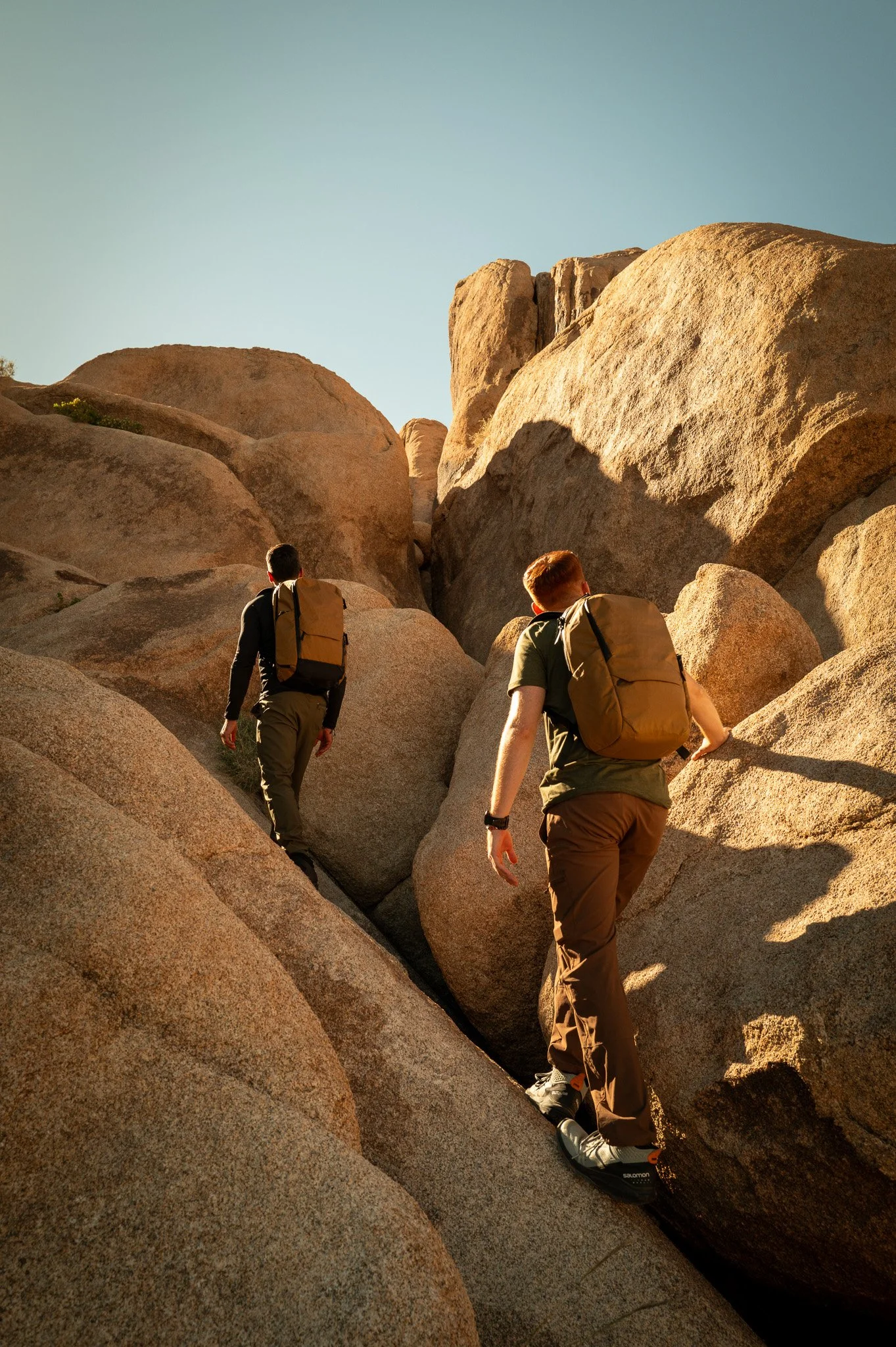 Two hikers with backpacks walking up a rocky, nature trail with large boulders, under a clear blue sky.