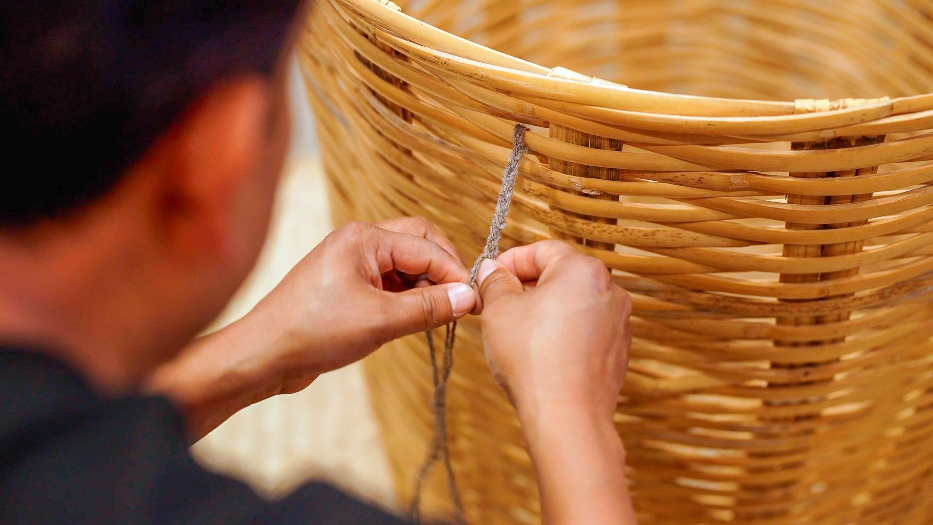 A person working on weaving a large wicker basket, threading a piece of natural fiber through the basket's weave.