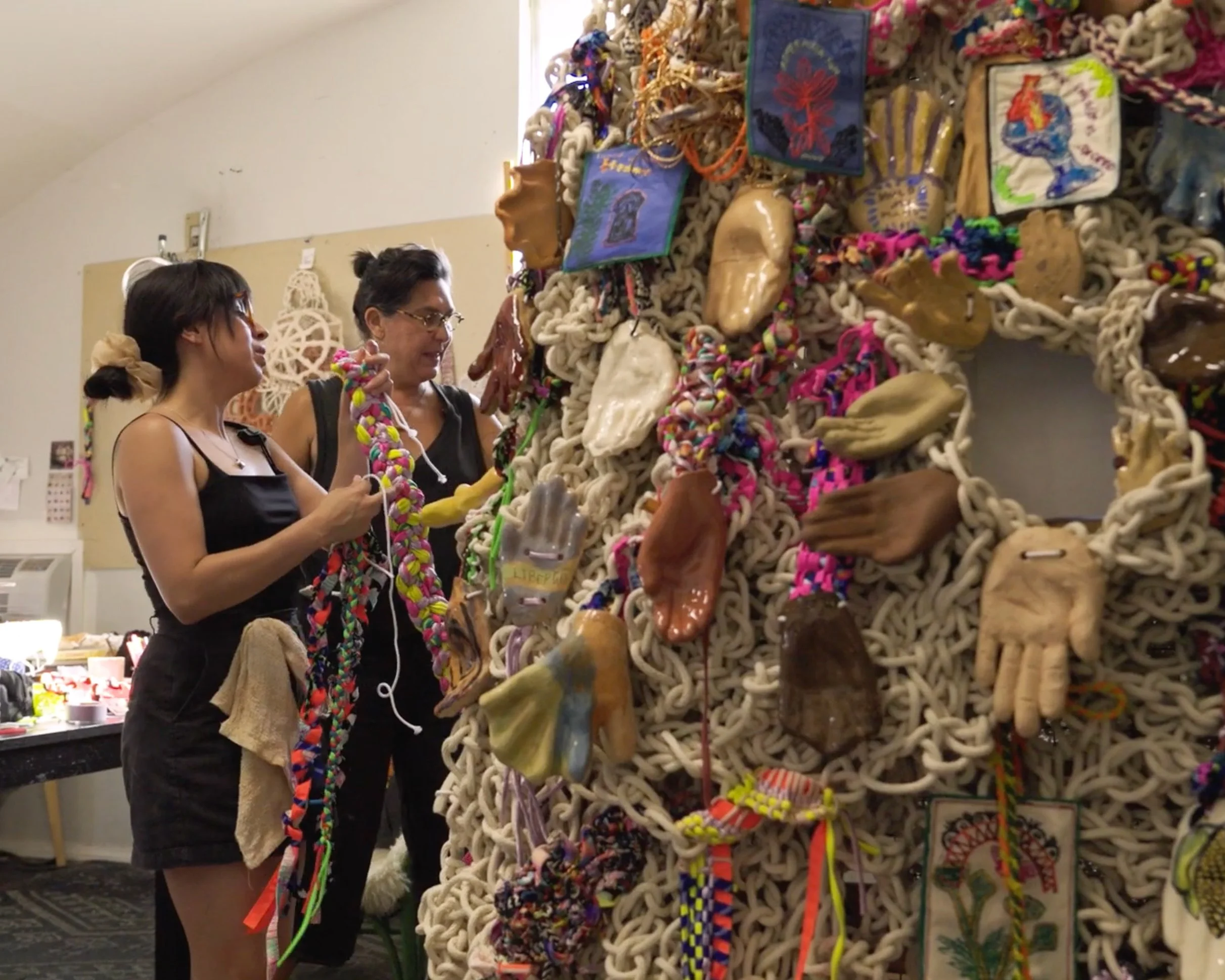 Two women looking at a large, colorful, woven art piece decorated with various textiles, beads, and small sculptures inside a room.
