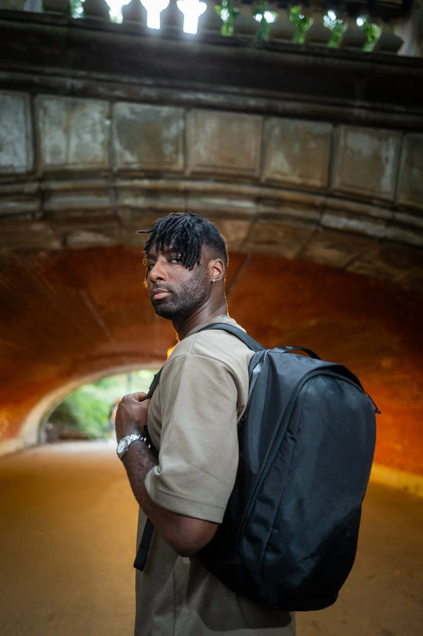 A man with dark hair styled in dreadlocks, wearing a beige shirt and carrying a black backpack, is standing under a bridge or tunnel with a brick arch and greenery visible in the background.