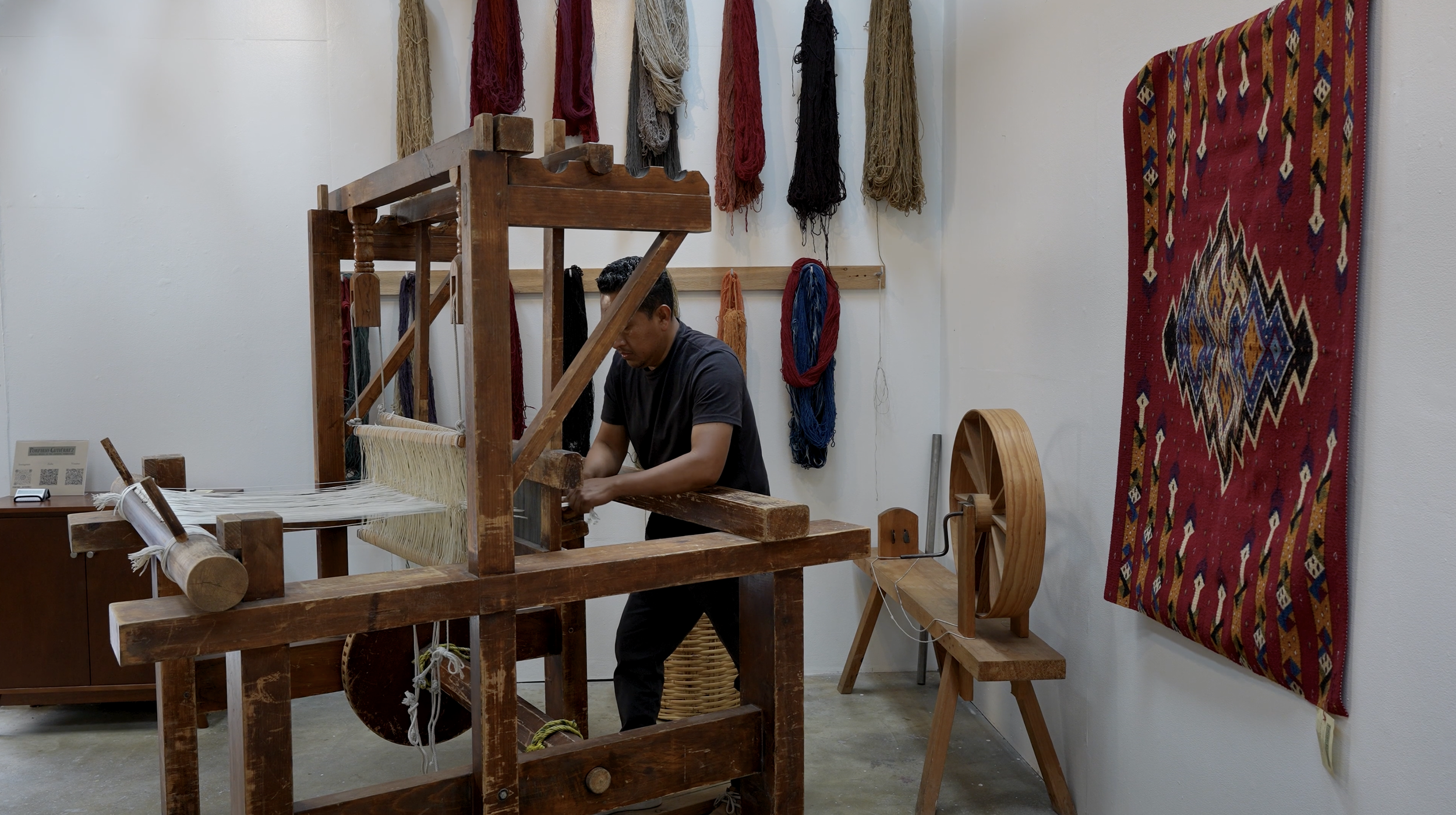 A man working on a wooden loom in a room decorated with colorful woven textiles and wall hangings.