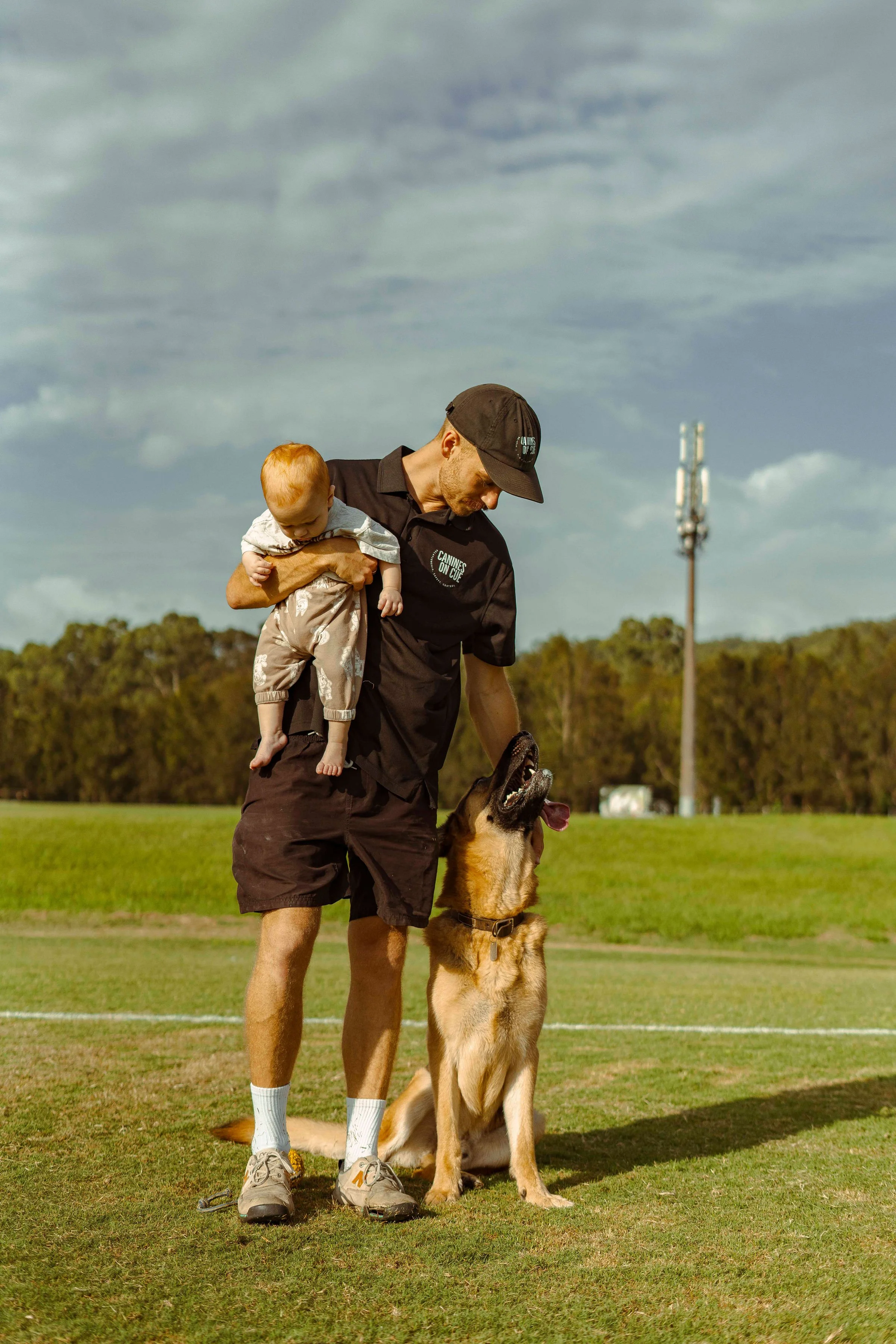 A man holding a young child on his shoulder and a German Shepherd dog sitting next to him on a grassy field during daytime.