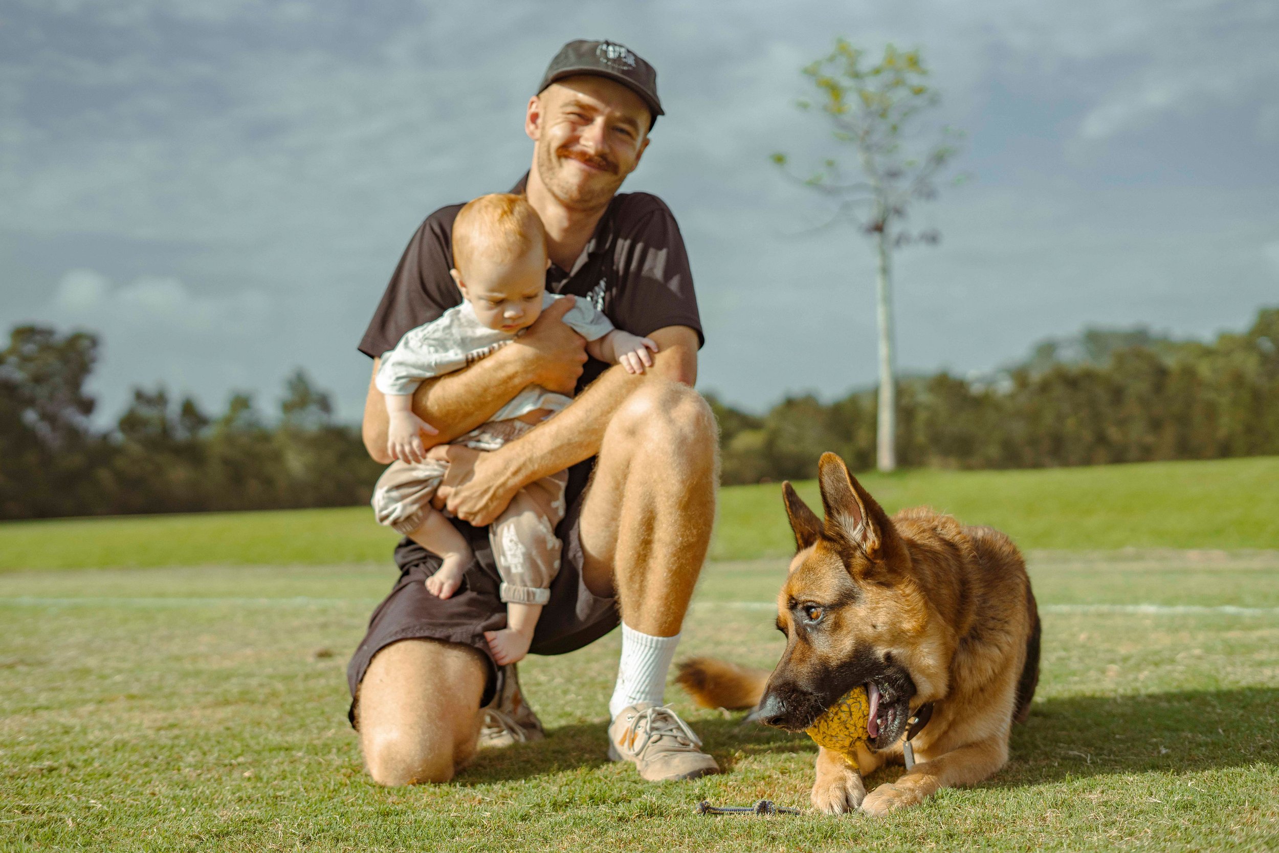 A man kneeling on the grass holding a young child, with a German Shepherd dog playing with a yellow ball nearby in an outdoor park setting with trees and a cloudy sky.