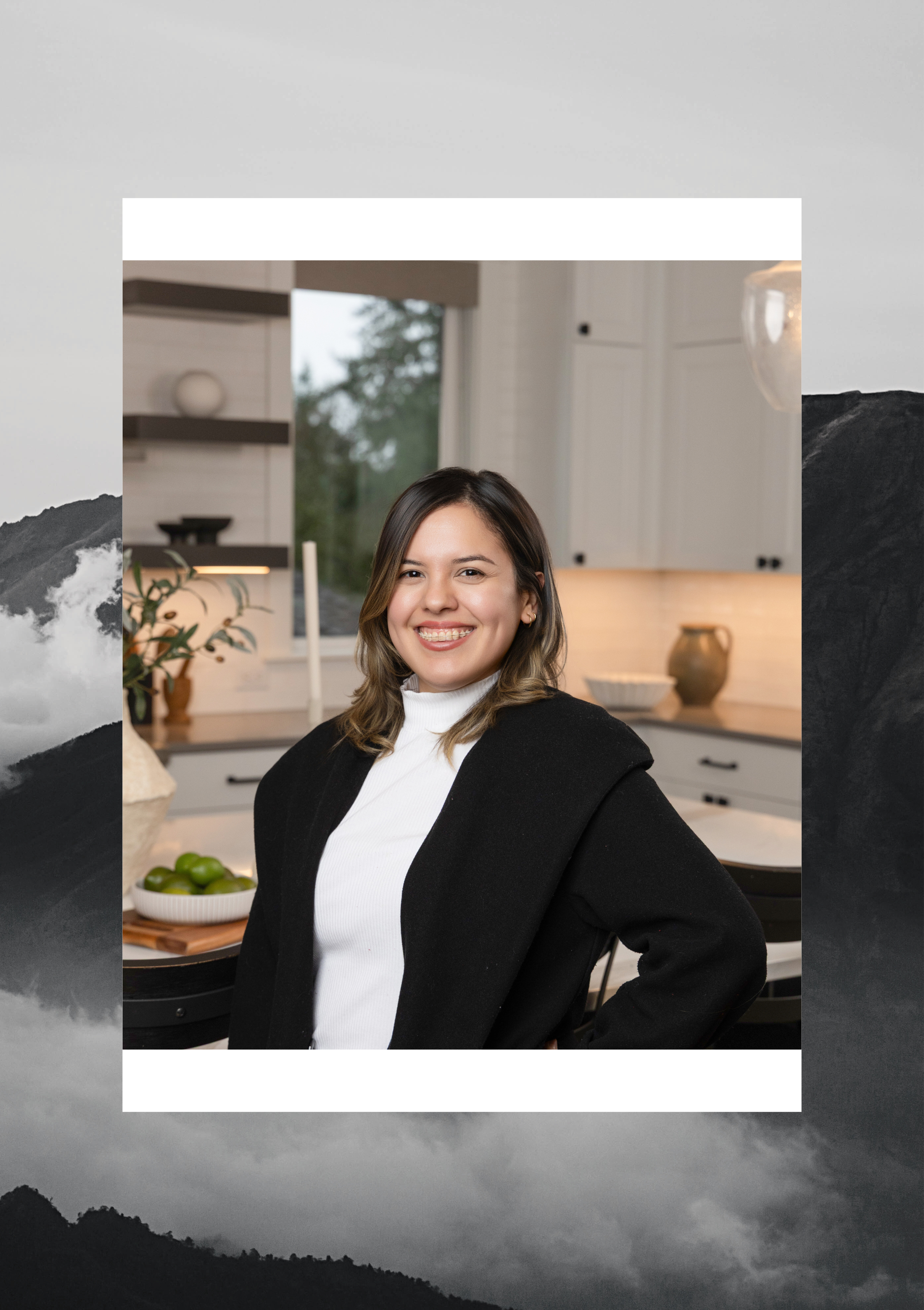 A young woman with shoulder-length brown hair, smiling, wearing a white turtleneck and a black coat, standing in a modern kitchen.