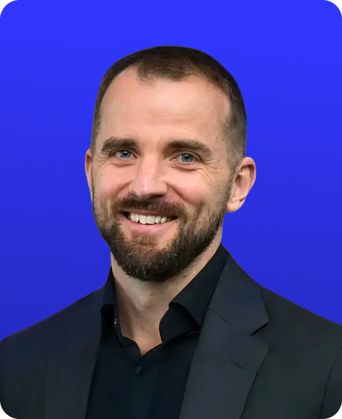 Portrait of a smiling man with short brown hair and a beard, wearing a dark suit and black shirt, against a blue background.