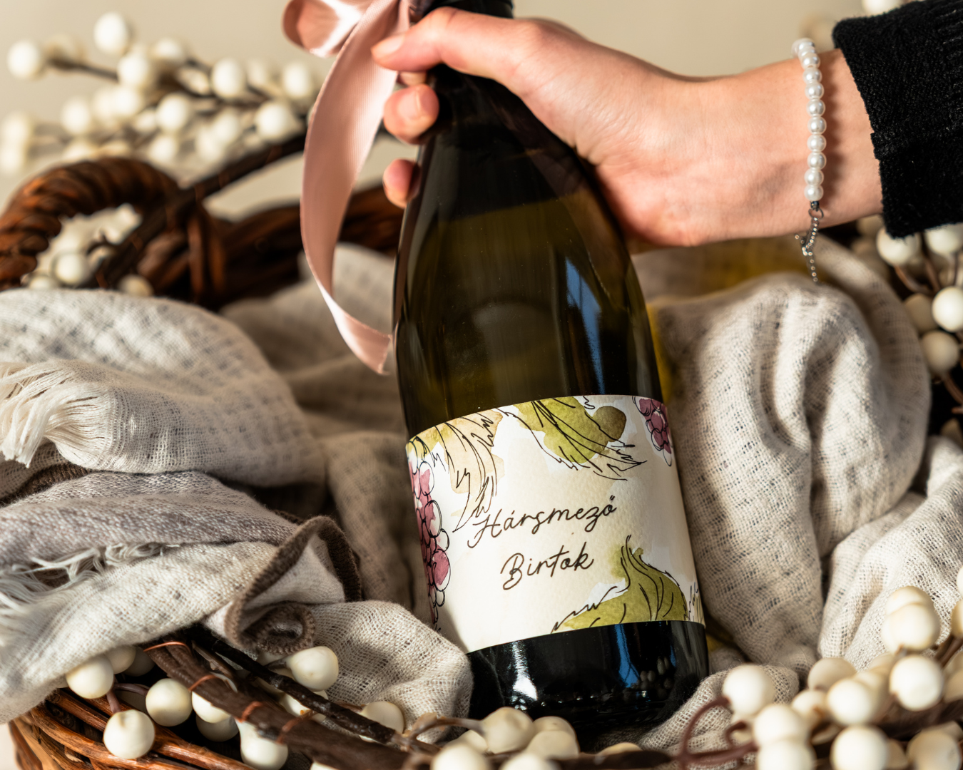 A hand holding a wine bottle with a decorative painted label, resting in a basket surrounded by white beads and soft fabric.