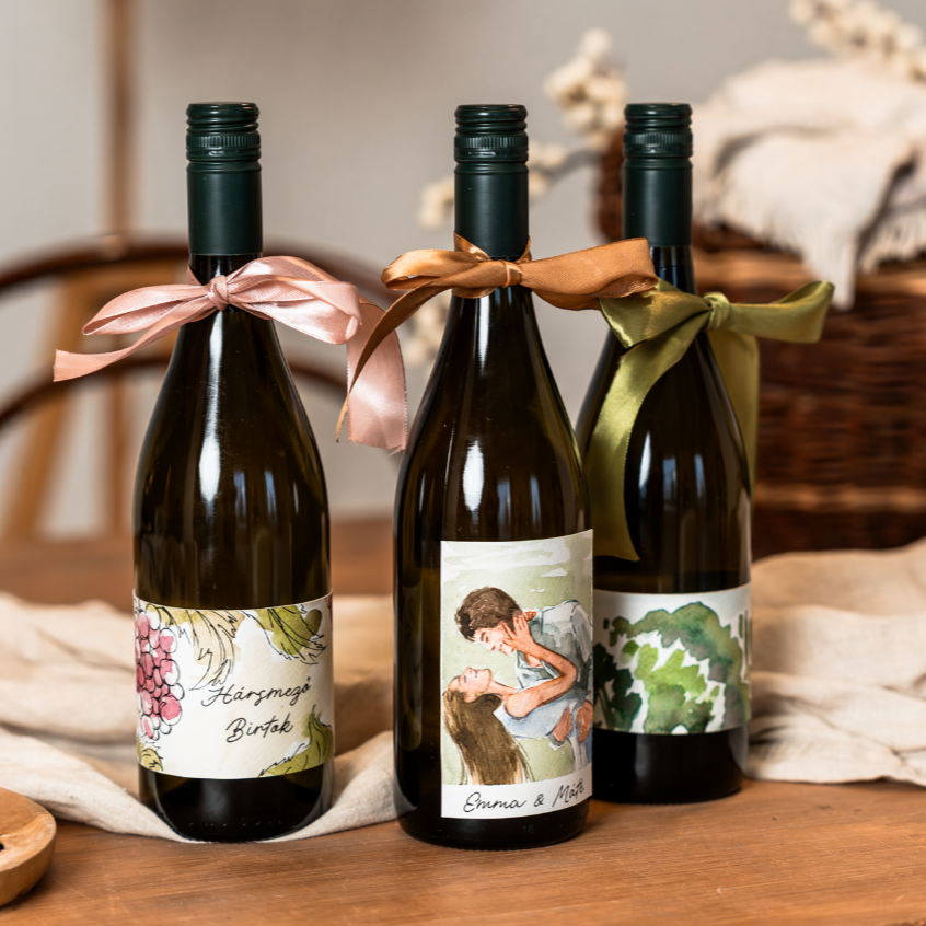 Three bottles of wine with personalized and painted labels and colored ribbons on the necks, placed on a wooden table with a beige cloth and a basket in the background.