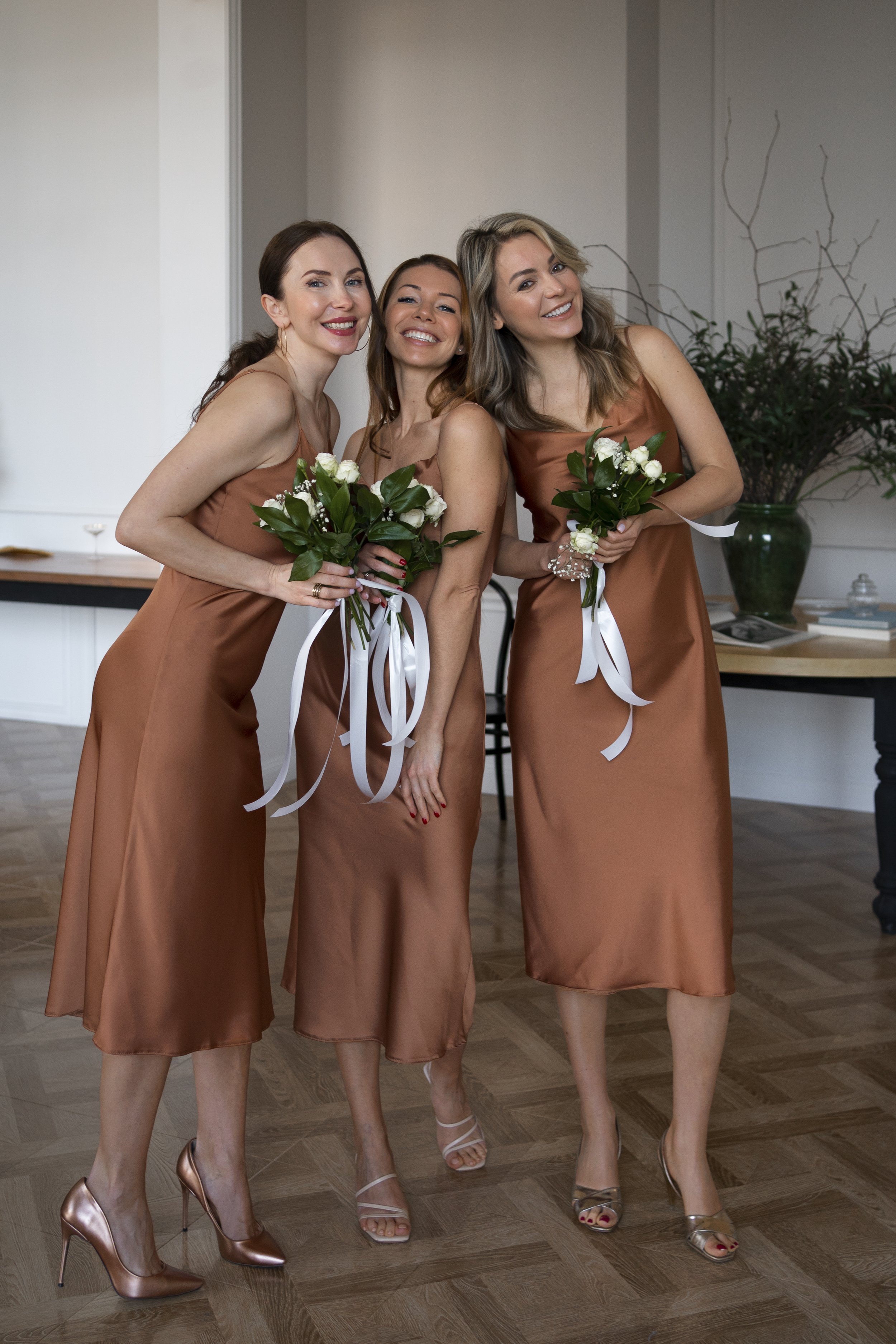 Three women in matching bronze satin dresses holding bouquets of white flowers, smiling and taking a selfie indoors.