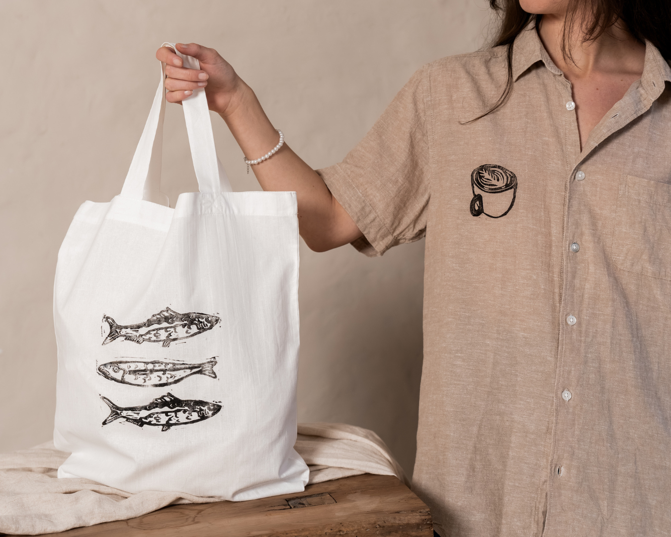 Person holding a white canvas tote bag with fish illustrations made with lino and lino ink, wearing a beige shirt with a coffee cup patch, and standing next to a wooden table.