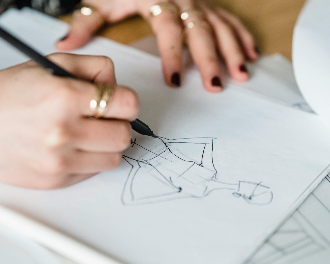 Close-up of a person's hand drawing on a sheet of paper, with another hand resting on the paper, on a wooden table.