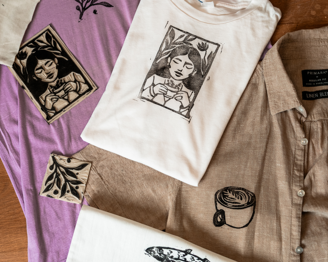 Assorted clothing and art pieces arranged on a wooden surface, including a white t-shirt with a black outline drawing of a woman holding a plant, a beige linen shirt with a black coffee mug illustration, and several small paintings or prints with black and white floral and abstract designs, some on fabric and some on paper.