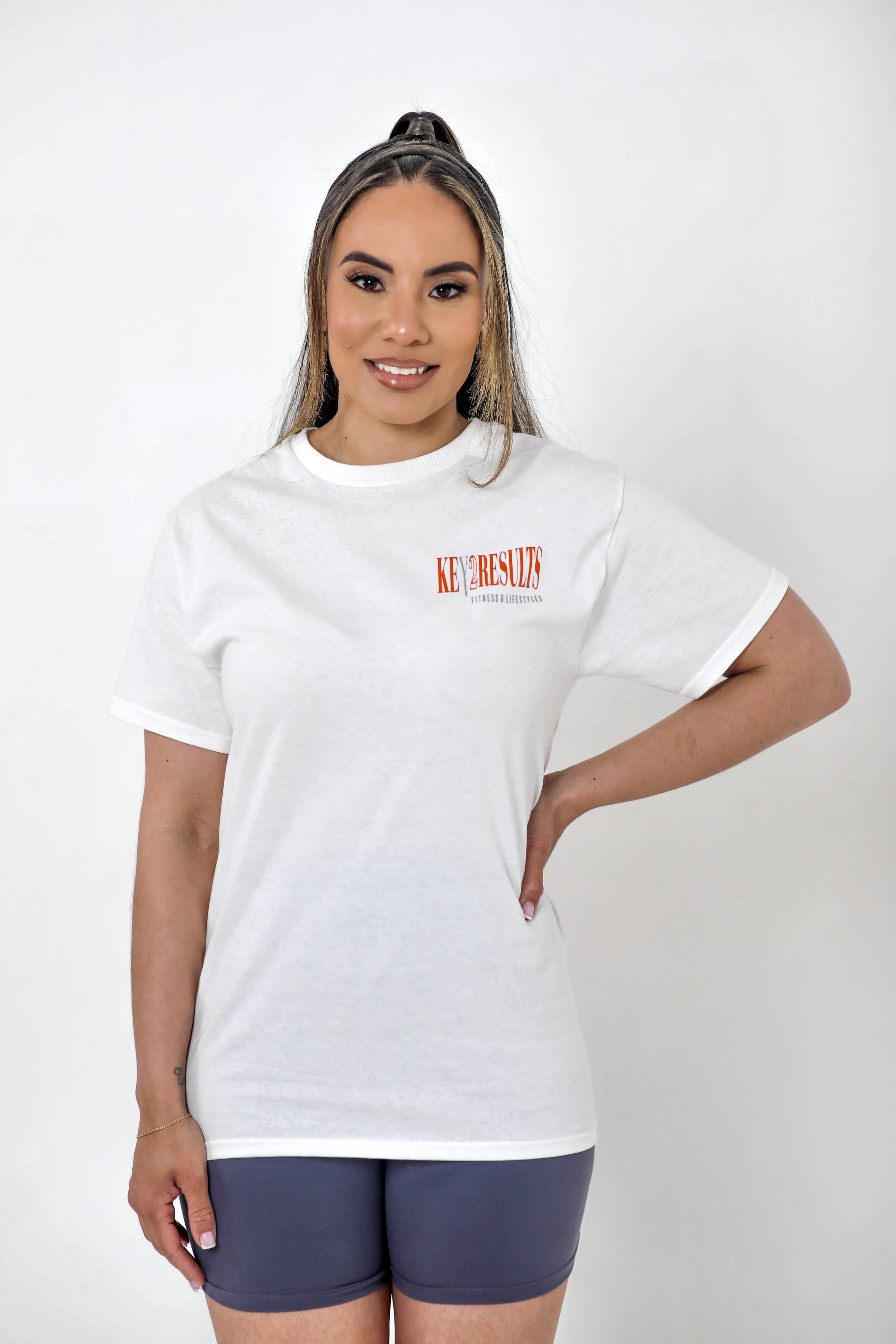 Young woman with long hair in a high ponytail, wearing a white T-shirt with red and black text, and dark gray shorts, standing against a plain white background.