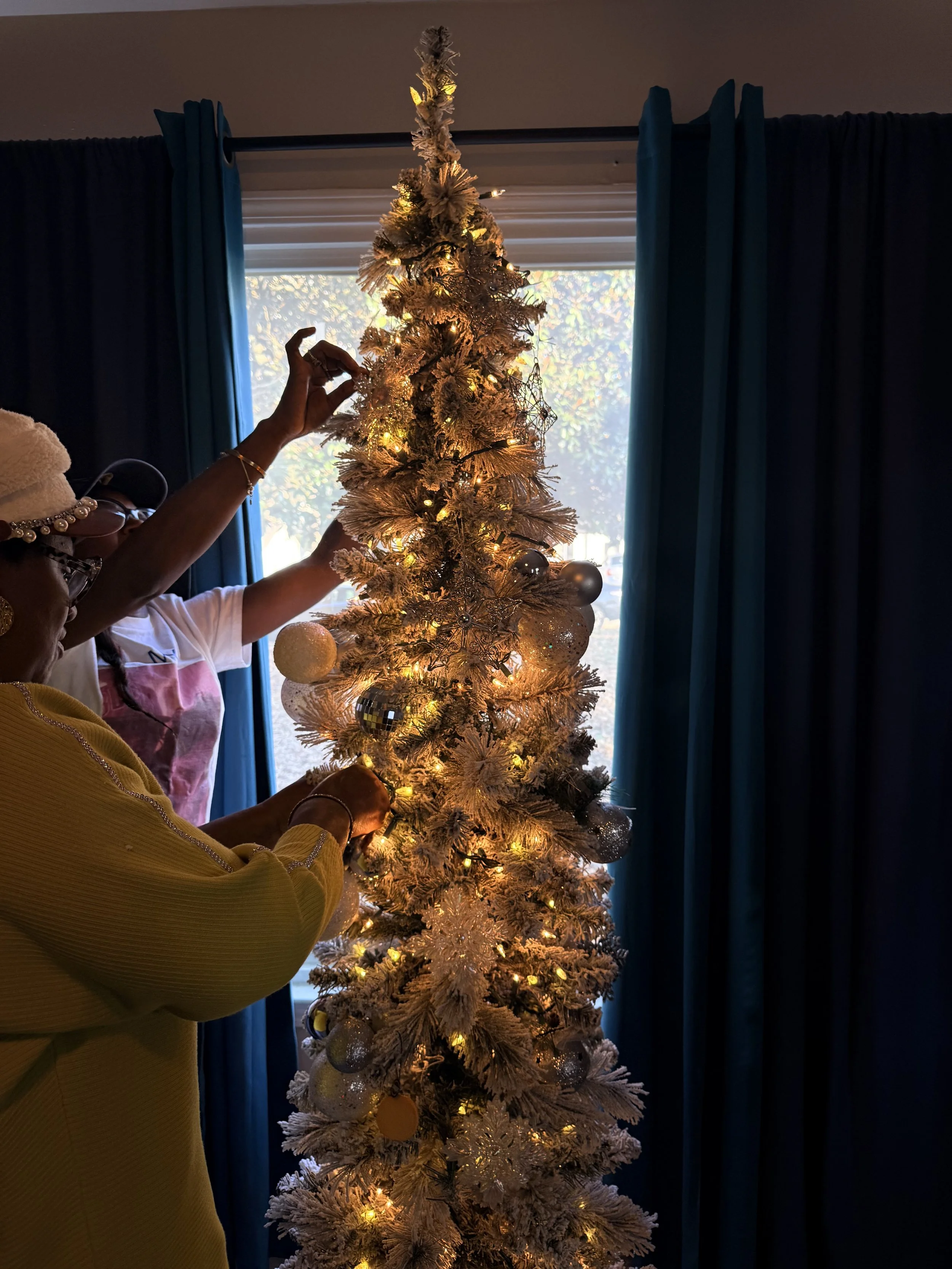 People decorating a white Christmas tree with white and silver ornaments and yellow lights in front of window with blue curtains.