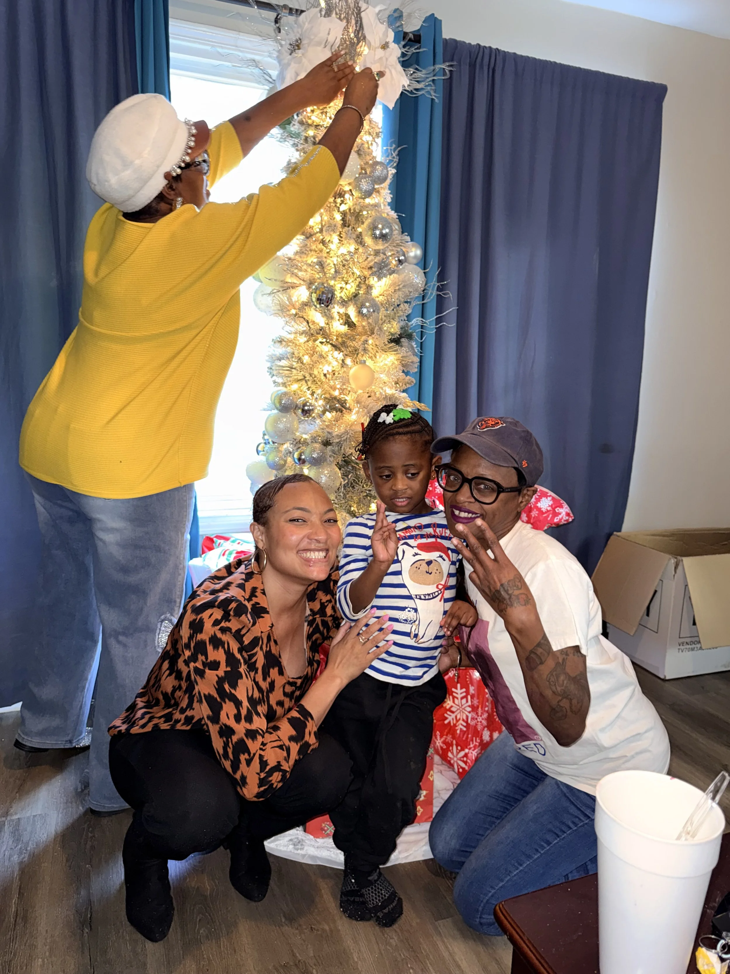 Four people, including a young girl, posing around a decorated Christmas tree, with one woman standing on a chair hanging a white ornament at the top, and a woman and a man kneeling beside the girl, smiling and making peace signs.