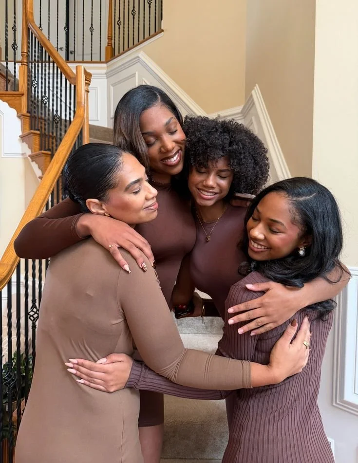 Four women hugging and smiling on a staircase inside a house.