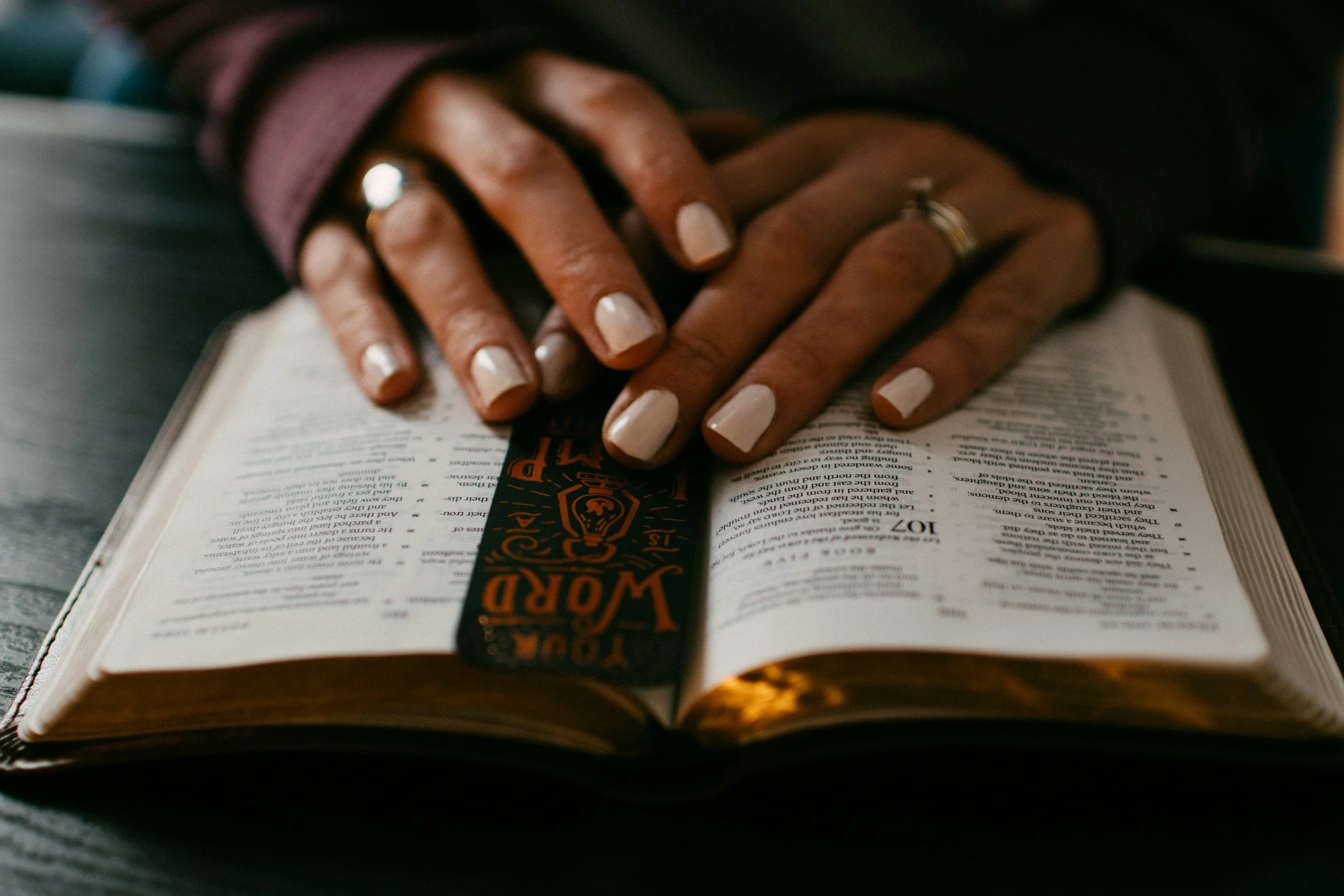 A person with manicured nails and rings on fingers reading a Bible, with a black and orange bookmark.