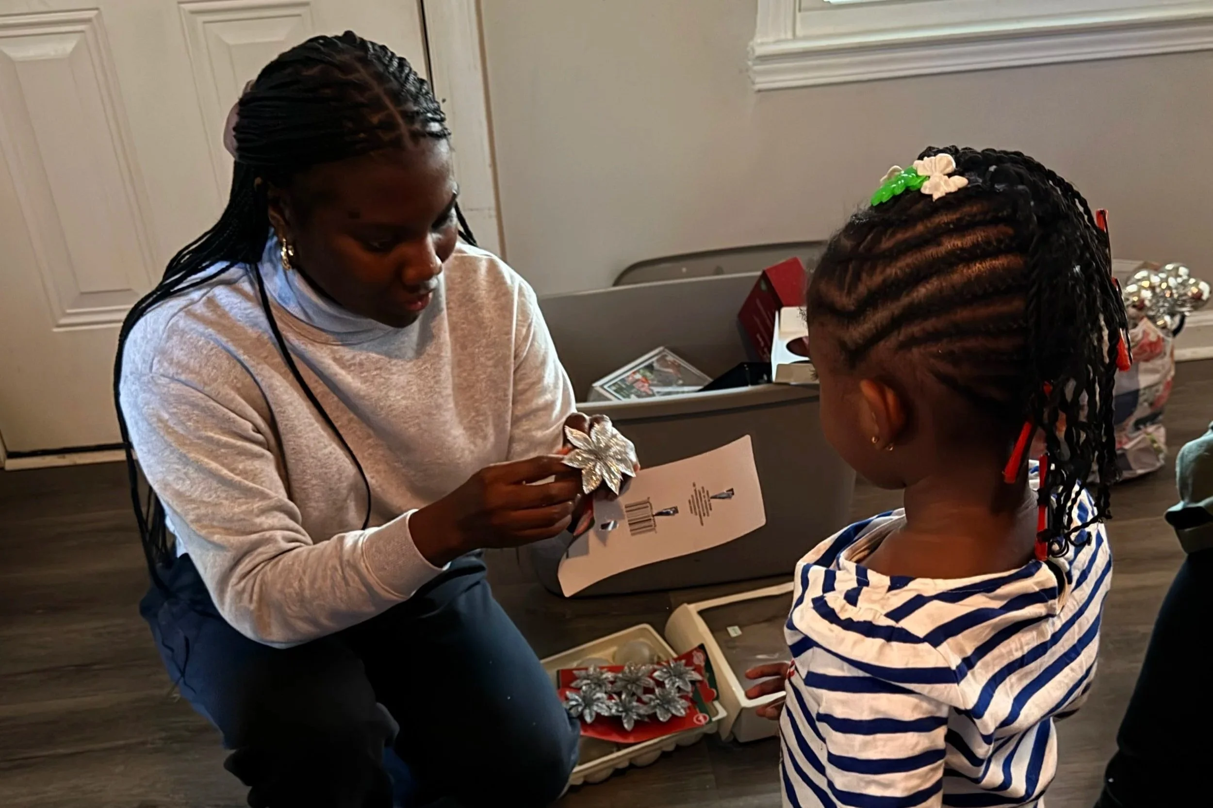 A young woman help a girl select a holiday decoration in a room, with gift boxes and Christmas decorations nearby.