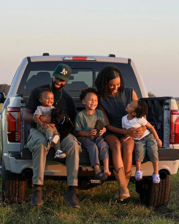 A family of four with two children and two dogs sitting on the back of a pickup truck outdoors during sunset, smiling and enjoying each other's company.