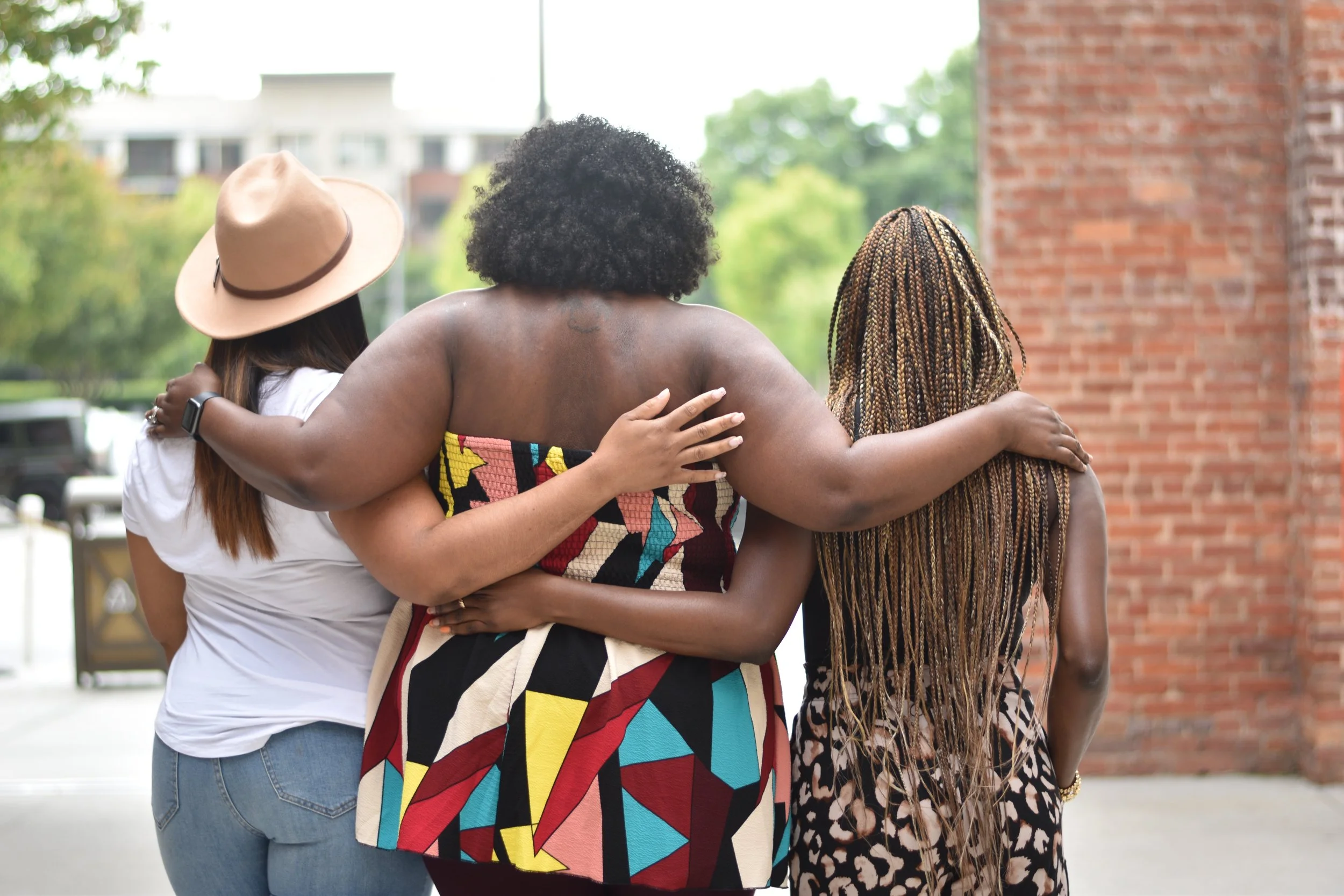 Three women standing outdoors with their backs to the camera, arms around each other, wearing diverse clothing and styles, near a brick wall.