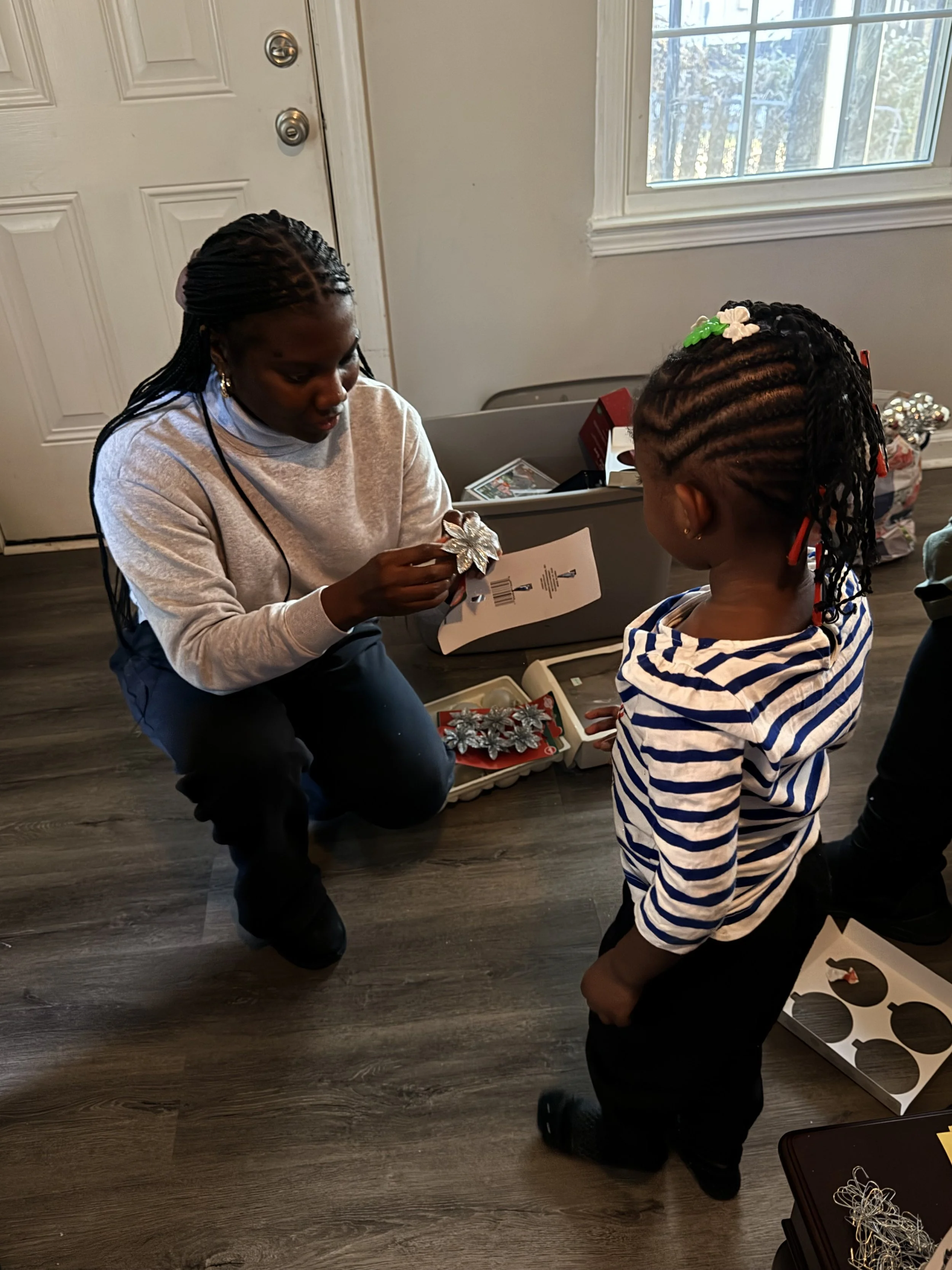 Woman showing a decorative flower item to a young girl indoors near a window, with a box of similar items on the floor.
