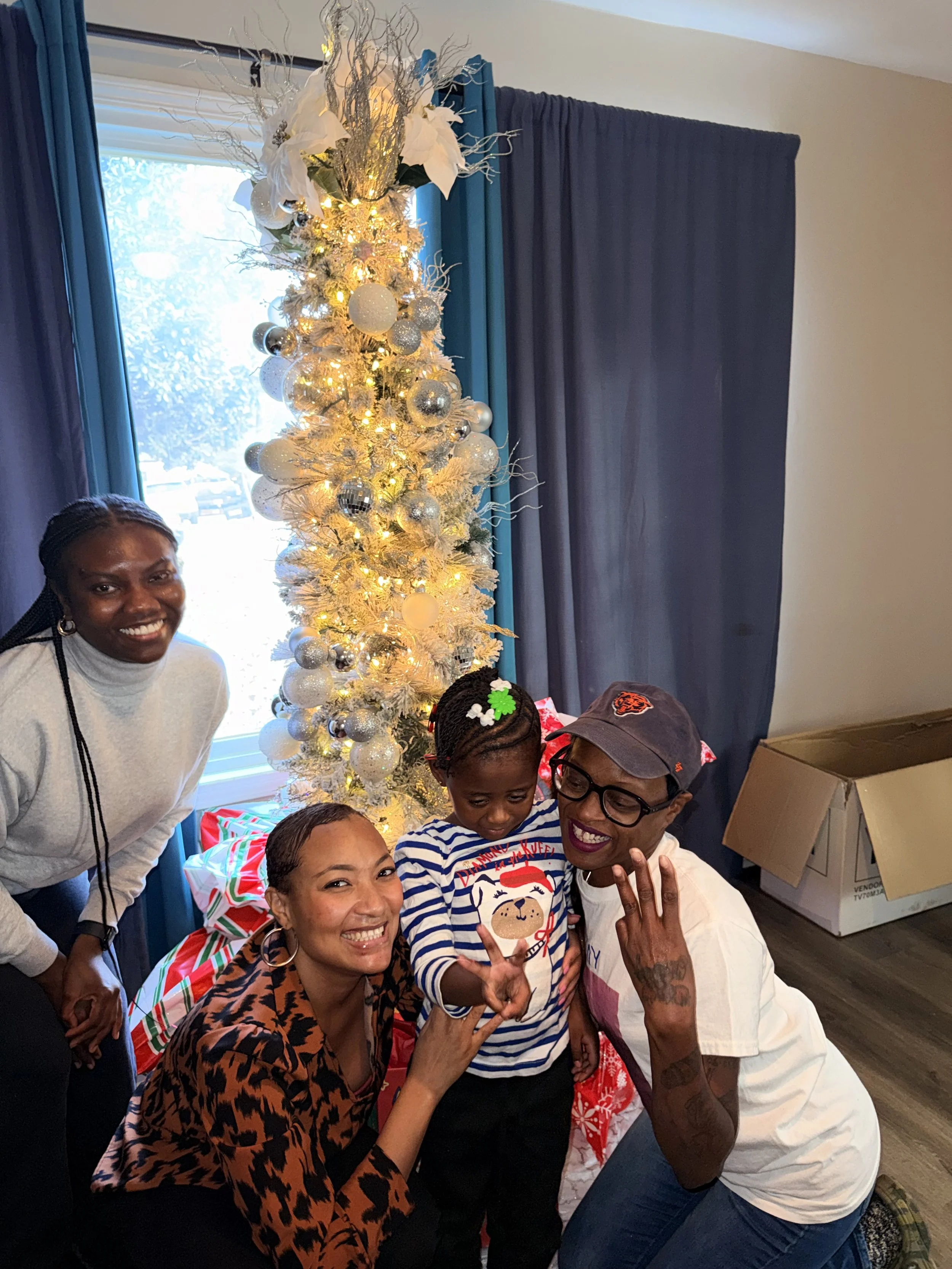 A group of four people, including a young girl, are gathered around a white Christmas tree decorated with silver and white ornaments, with a window and blue curtains in the background. They are smiling and posing for the photo inside a room.