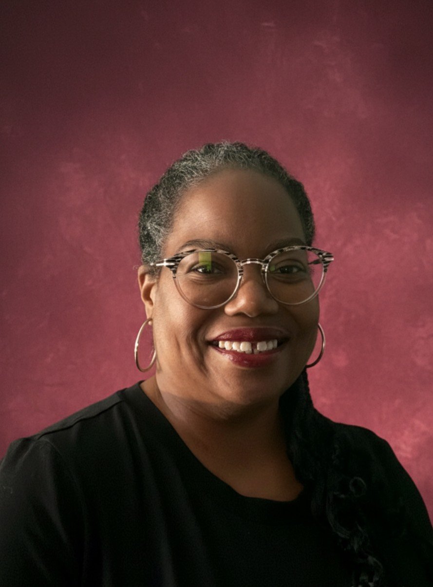 A woman with glasses and hoop earrings smiling in front of a pink textured background.
