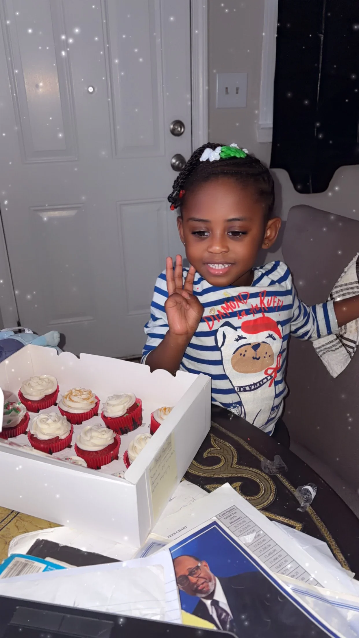 A young girl with braids and a colorful hair accessory making a peace sign next to a box of red cupcakes with white frosting in a living room.