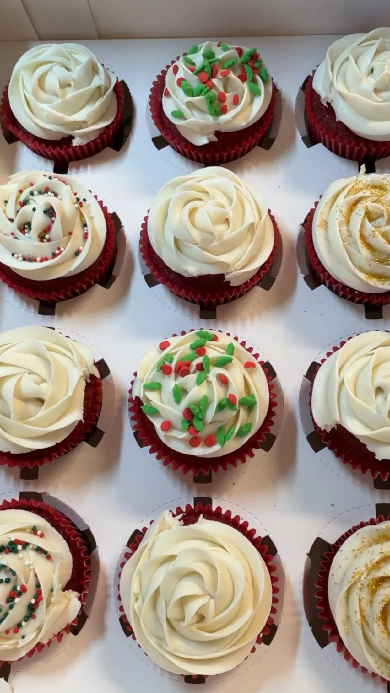 Assorted cupcakes with white frosting, some decorated with colorful sprinkles and others with gold or green decoration, arranged in a grid pattern in red cupcake liners.