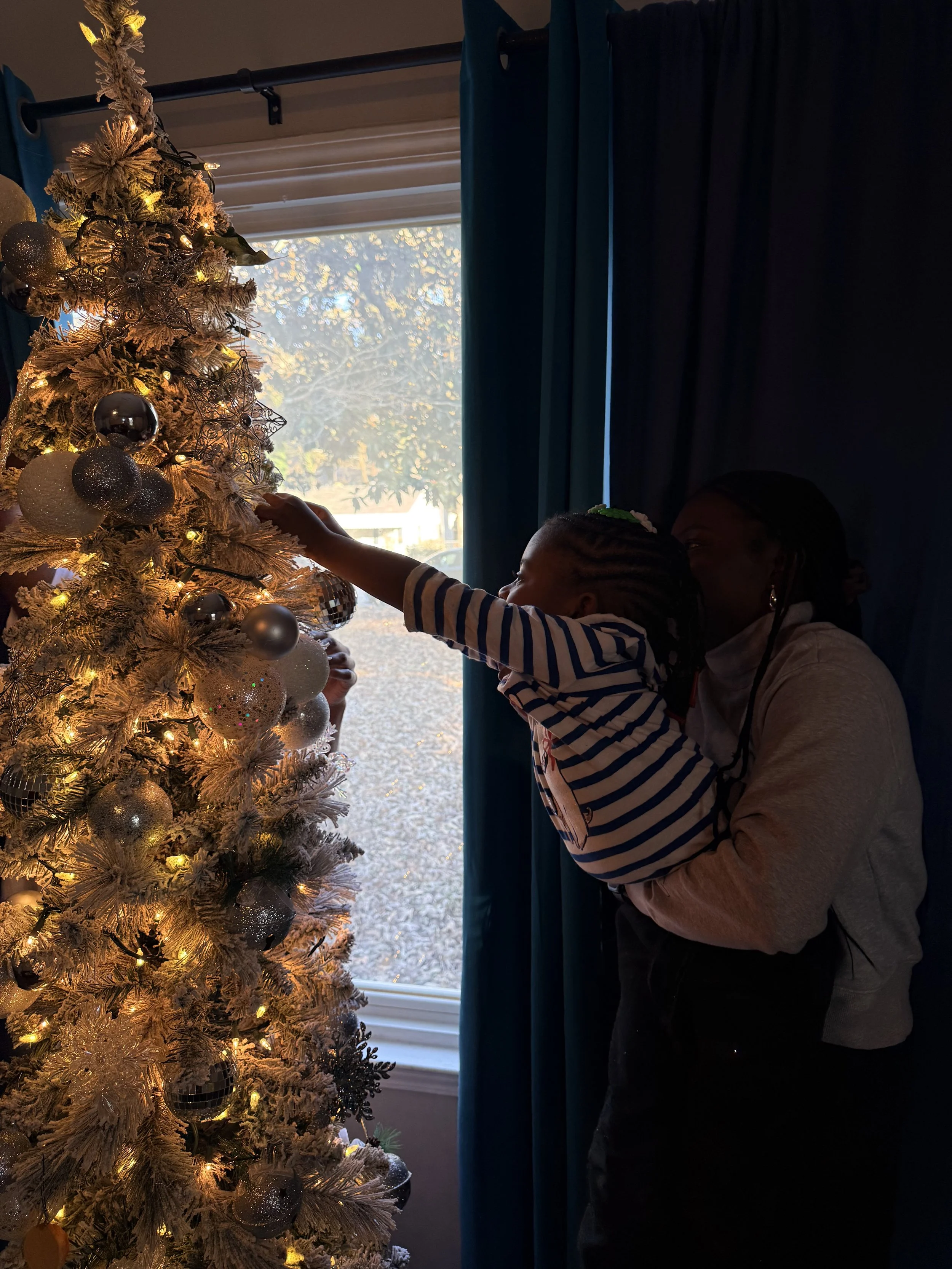 A child hanging ornaments on a decorated Christmas tree while a woman holds them, in front of a window with curtains.