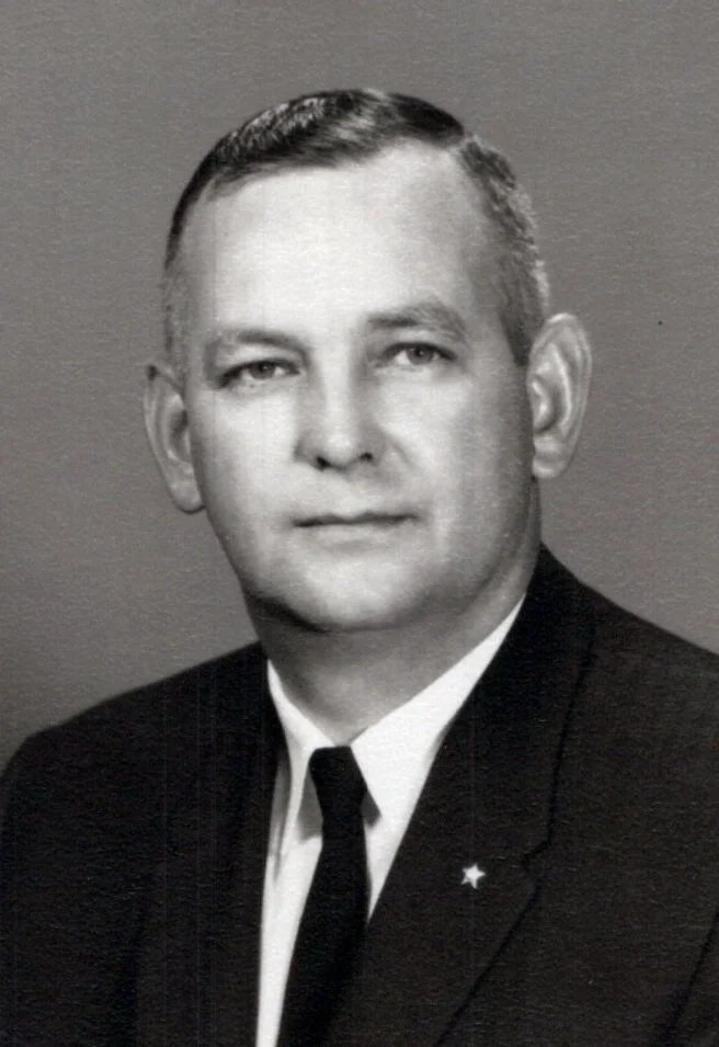 Black and white portrait of a man in a suit with a white shirt and dark tie, featuring short hair and a neutral expression.