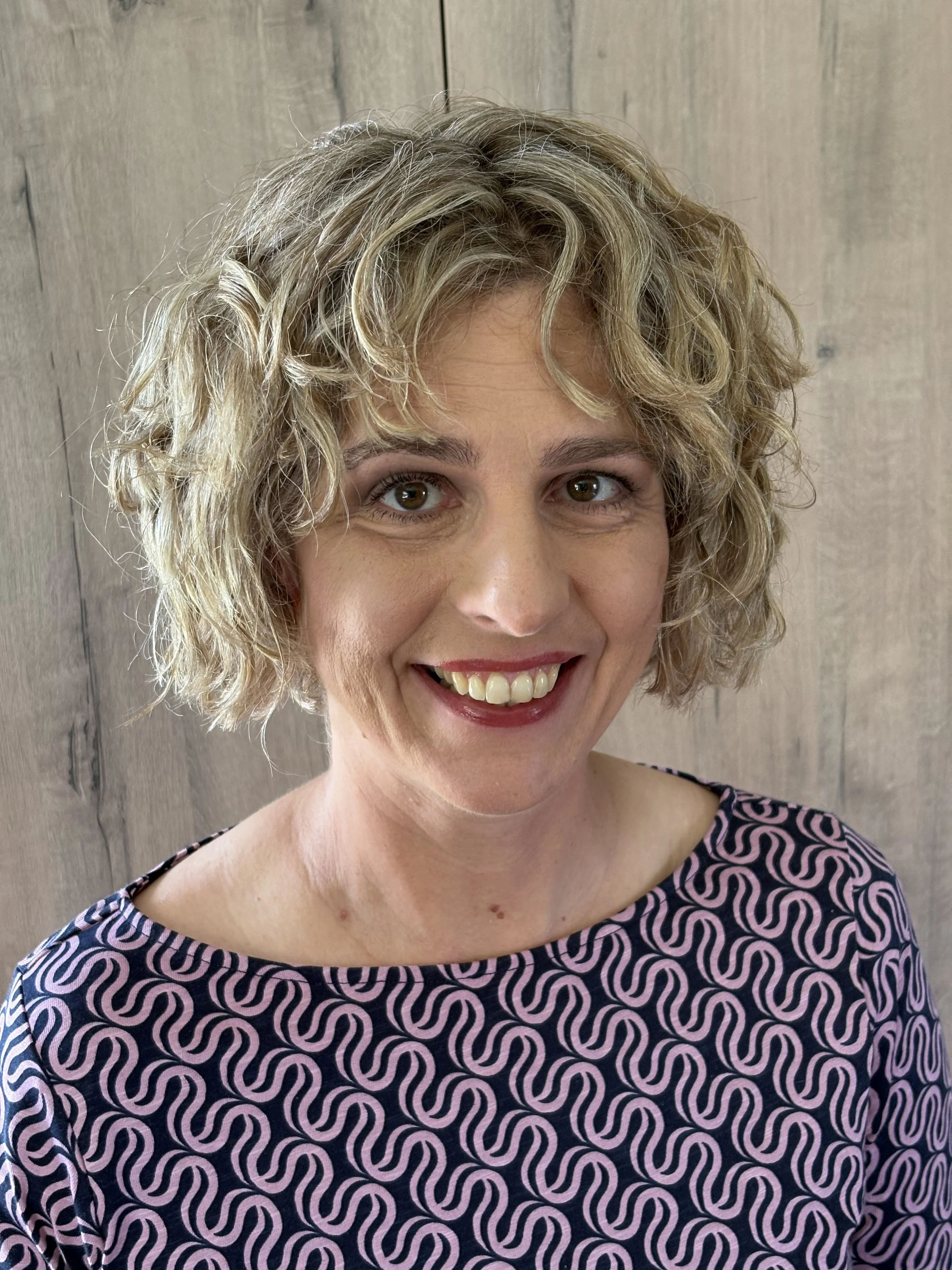 A woman with curly blonde hair smiling, wearing a patterned top with pink and black wavy lines, standing in front of a wooden background.