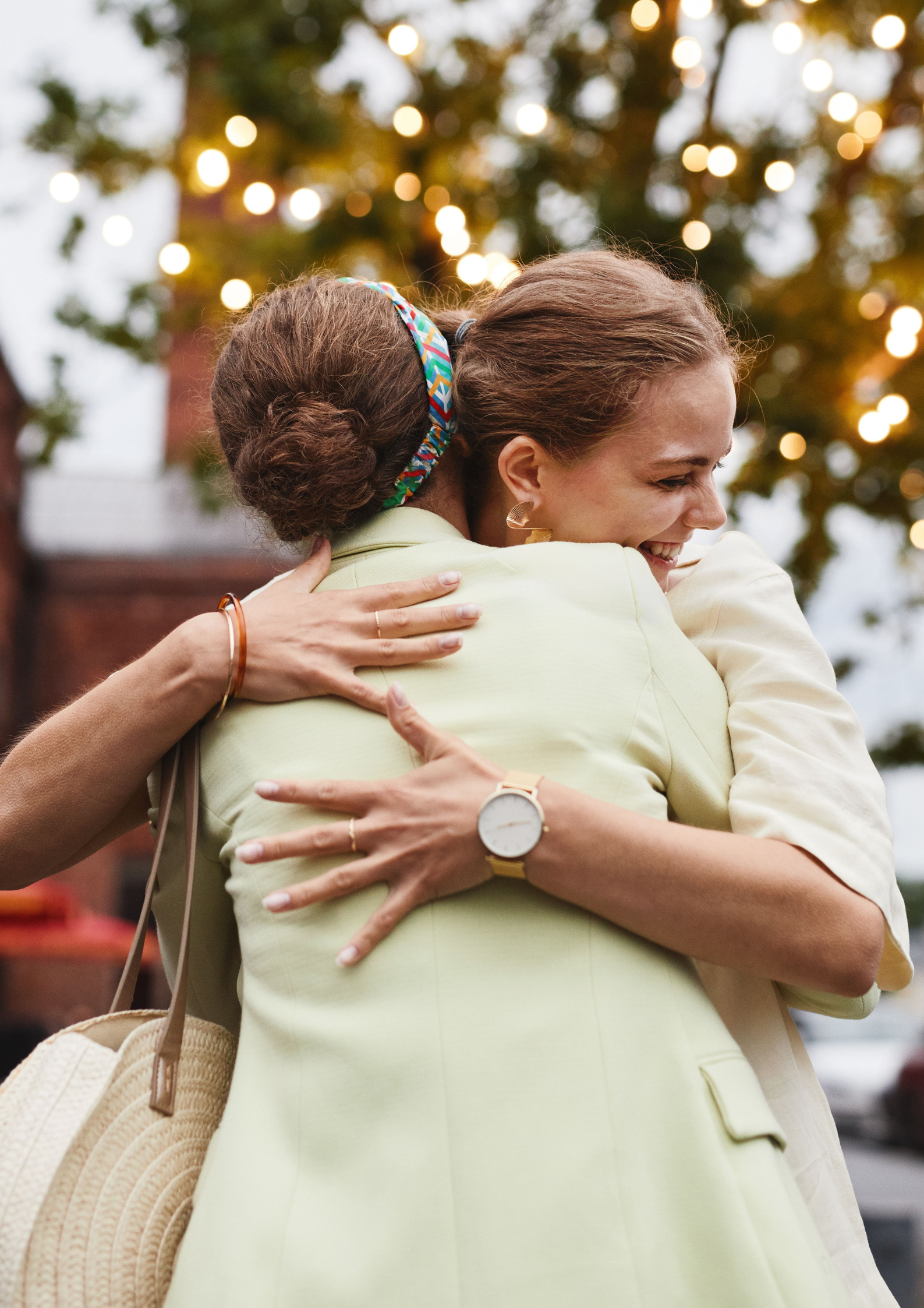Two women smiling and hugging in an outdoor setting during evening, with blurred lights and trees in the background.