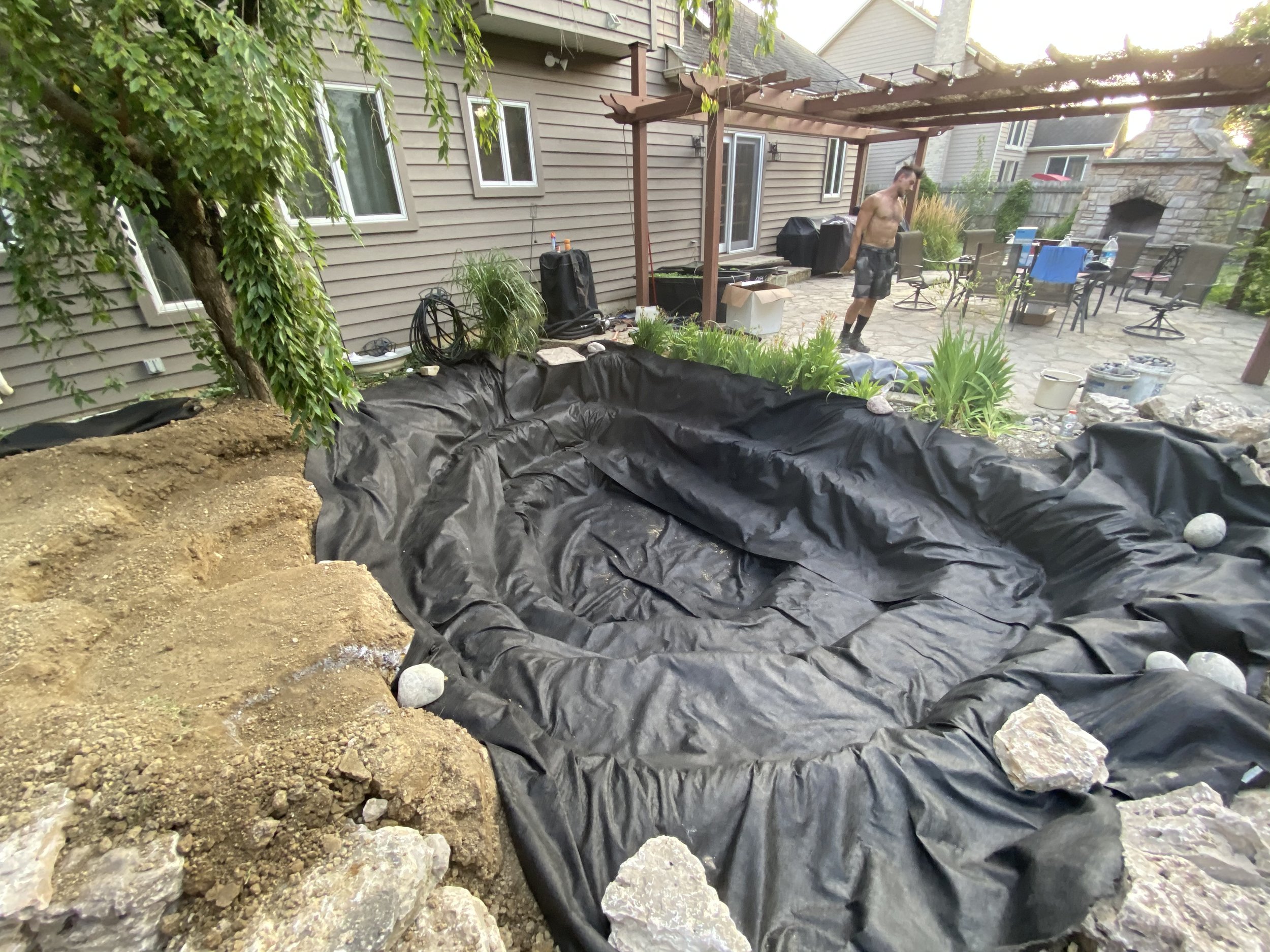 Backyard pond construction with black liner, rocks, and a pergola.